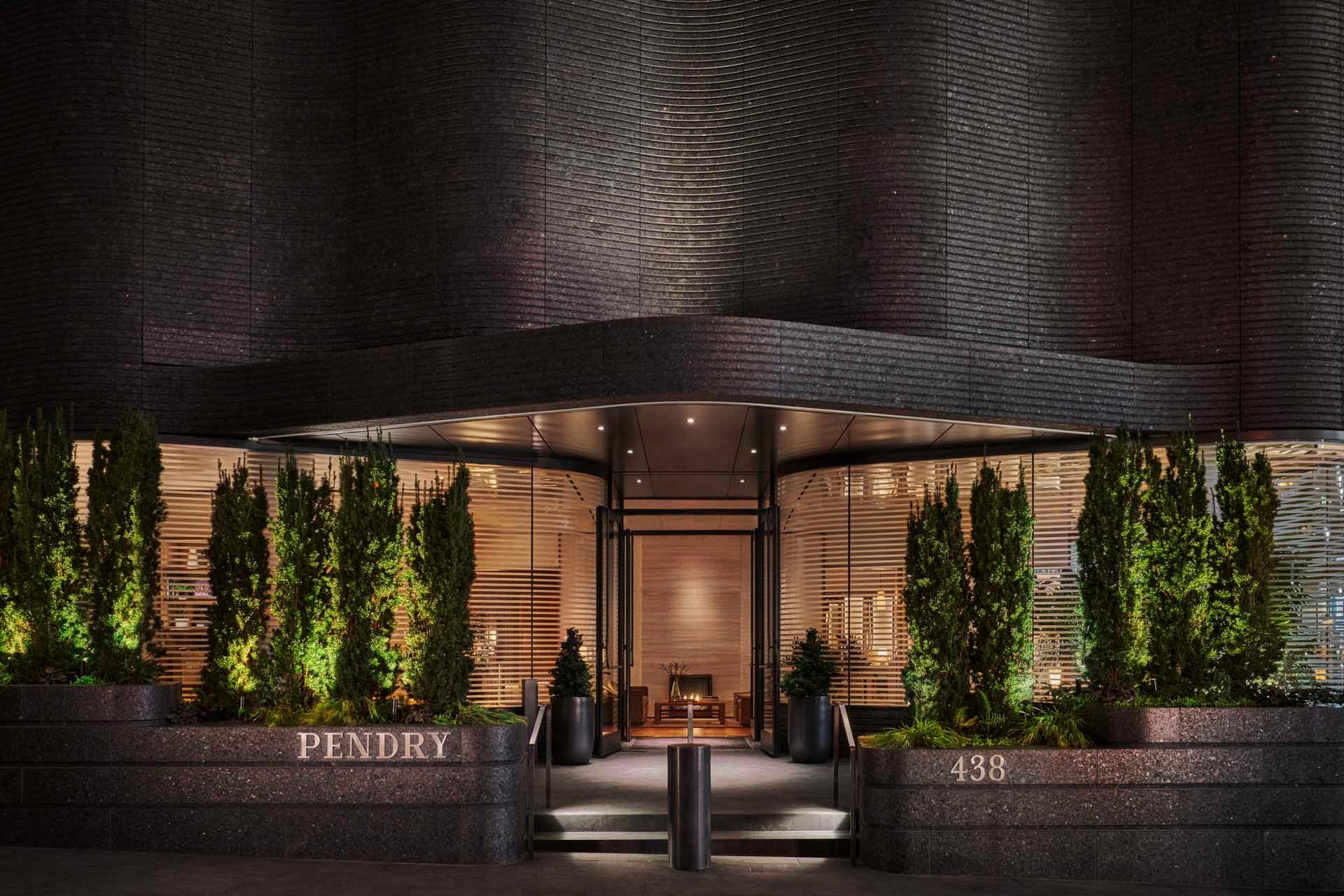 Front entrance of a modern building with curved architecture, featuring well-lit windows and vertical wood slats. Tall, neatly trimmed shrubs line the entrance, and the numbers "438" are visible. Elegant outdoor lighting enhances the facade.