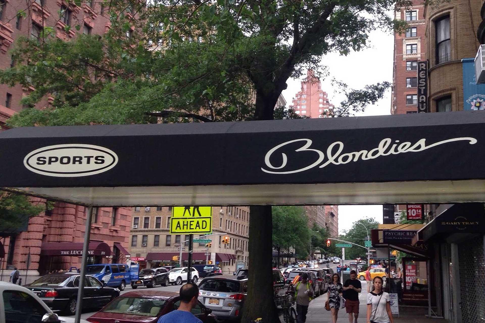 A black awning with "SPORTS Blondies" written in white hangs over a busy city sidewalk with pedestrians, parked cars, trees, and tall brick buildings in the background.