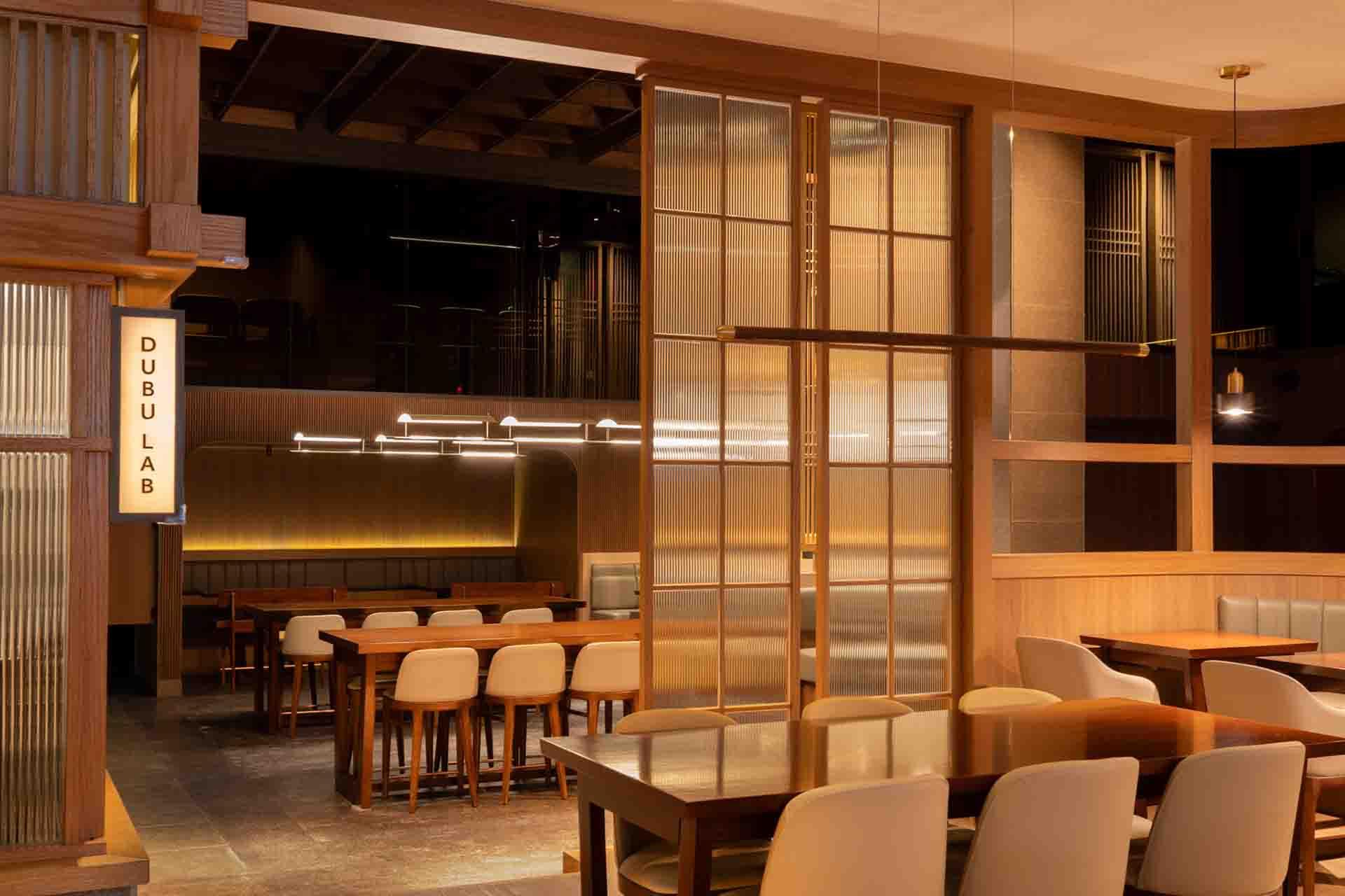 Modern restaurant interior with wooden tables and beige chairs, warm lighting, and a sign reading "DUBU LAB" on a wall divider. The space appears clean, organized, and inviting.