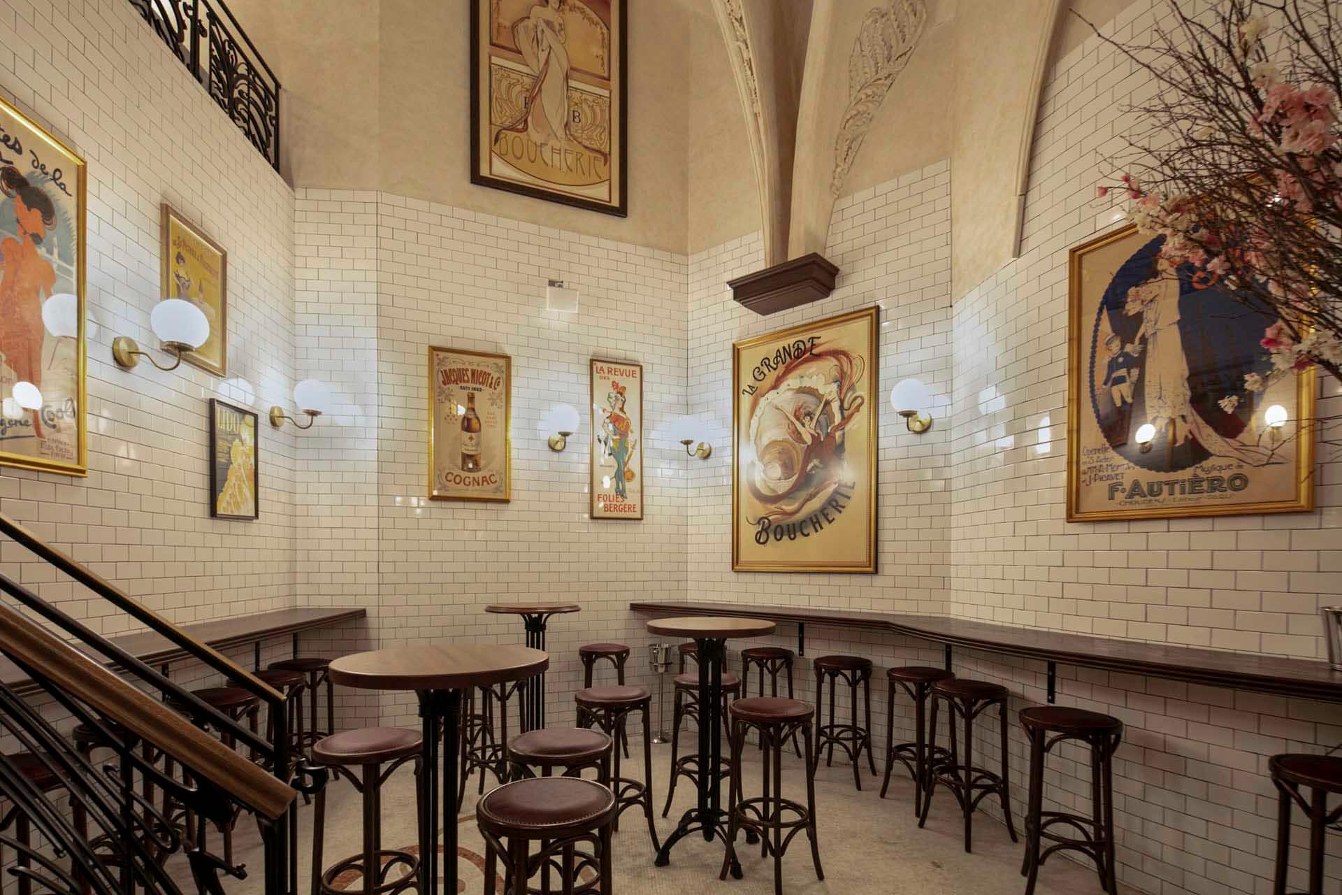 A vintage-style room with white tiled walls and art nouveau posters. The room features several tall, round tables and wooden stools. Ornate lighting fixtures hang on the walls, and a staircase with a black railing is visible on the left.