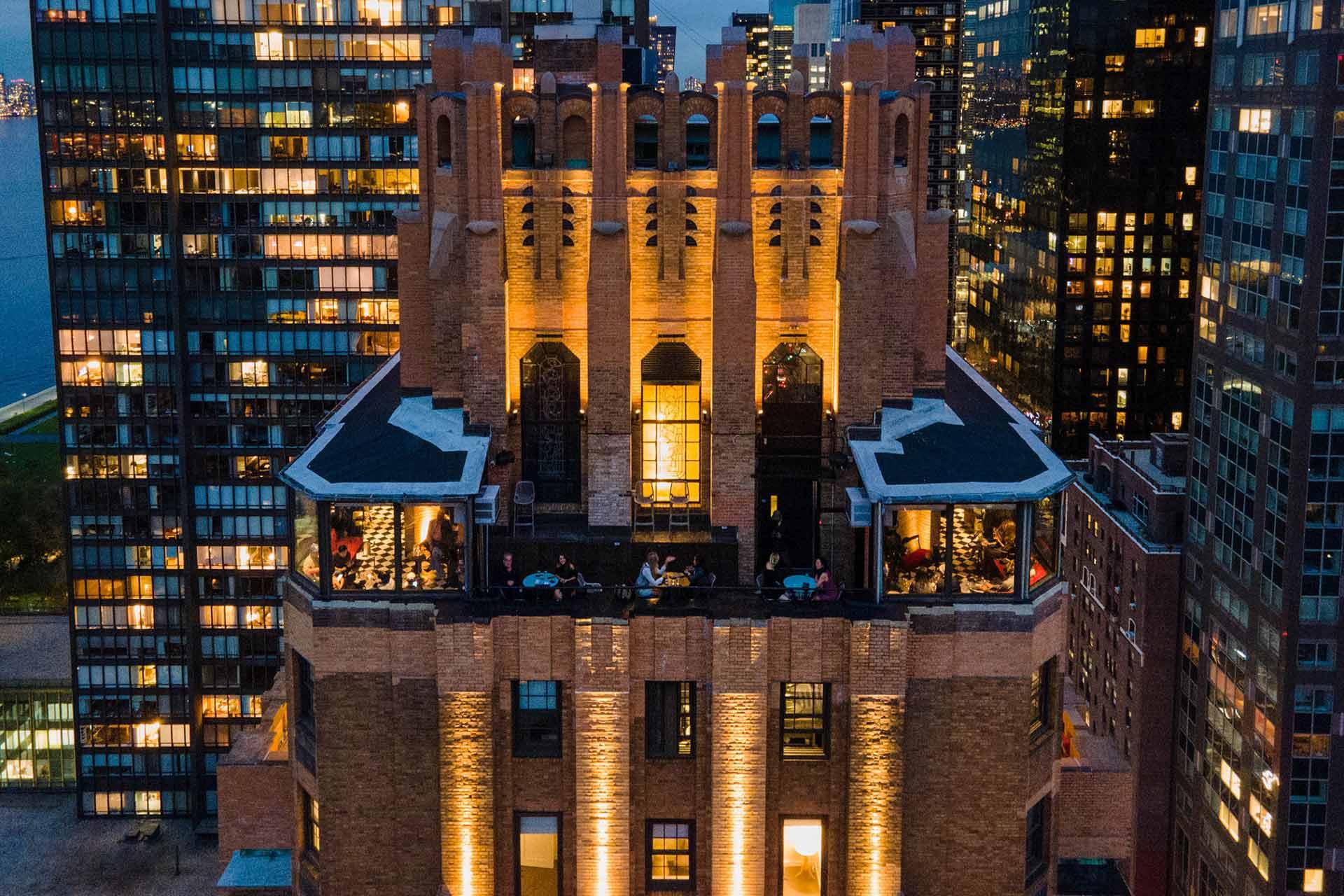 Aerial view of a tall, illuminated brick building with gothic architectural details, surrounded by modern glass skyscrapers at dusk. People are visible dining on terraces. City lights glow in the background.
