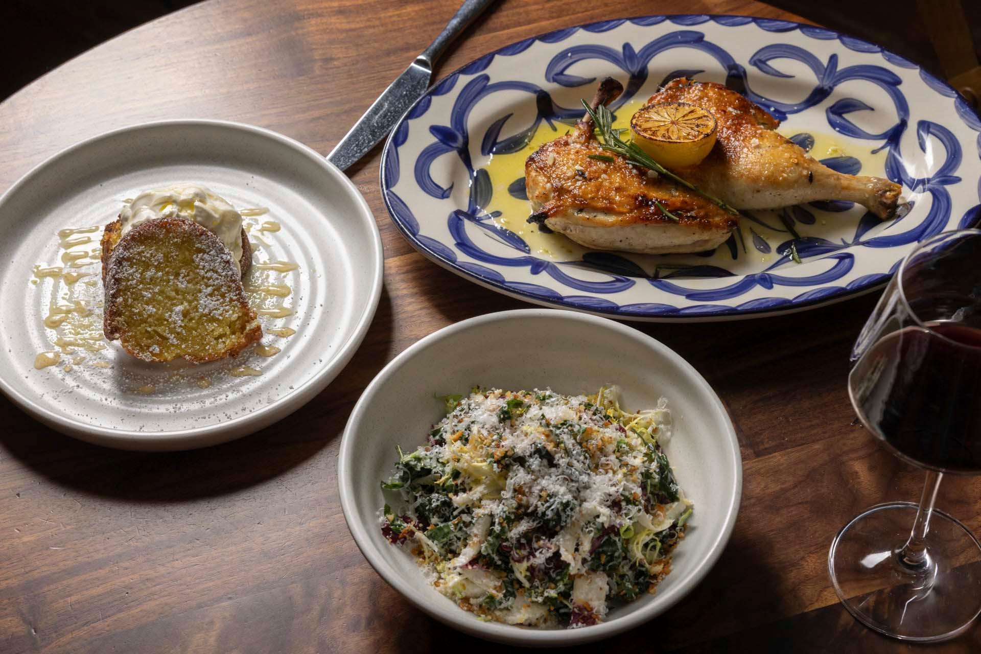 A wooden table set with a meal: a plate of grilled chicken with lemon garnish, a bowl of leafy green salad topped with grated cheese, a dessert cake with ice cream and drizzle, and a glass of red wine.