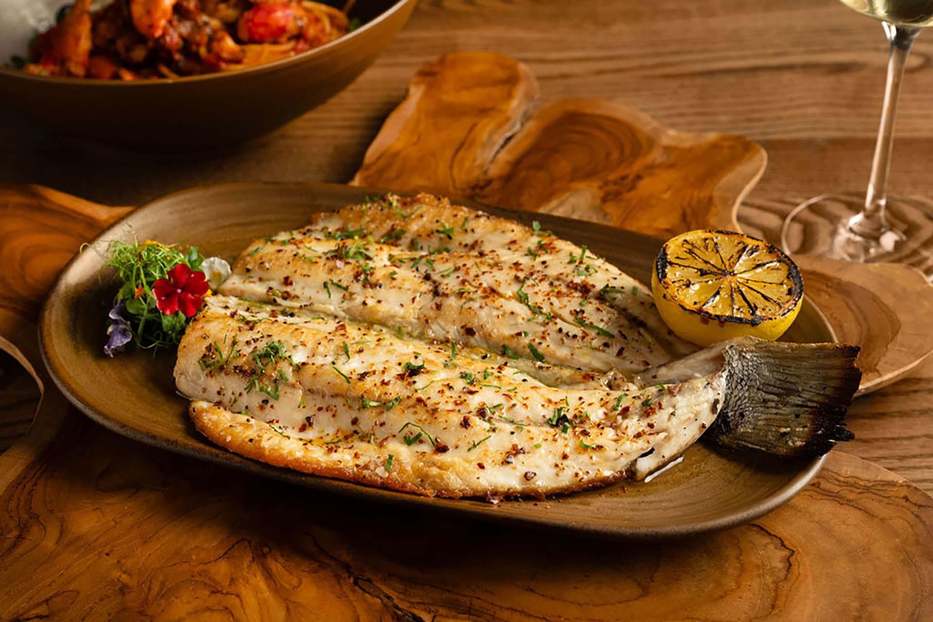Grilled whole fish served on a brown plate with herbs, a charred lemon half, and a small garnish; wooden table and a glass of white wine in the background.