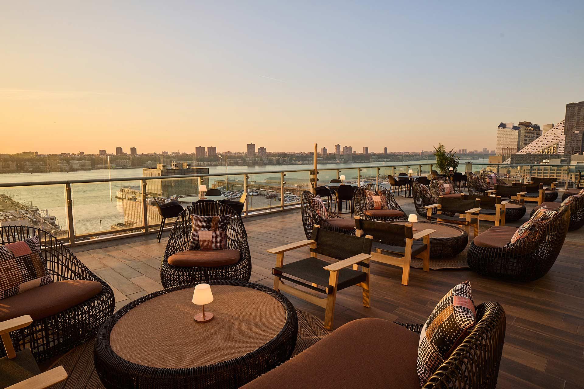 Rooftop lounge at sunset with wicker chairs, round tables, small lamps, and glass railing overlooking a river and city skyline. The atmosphere is relaxed and inviting.