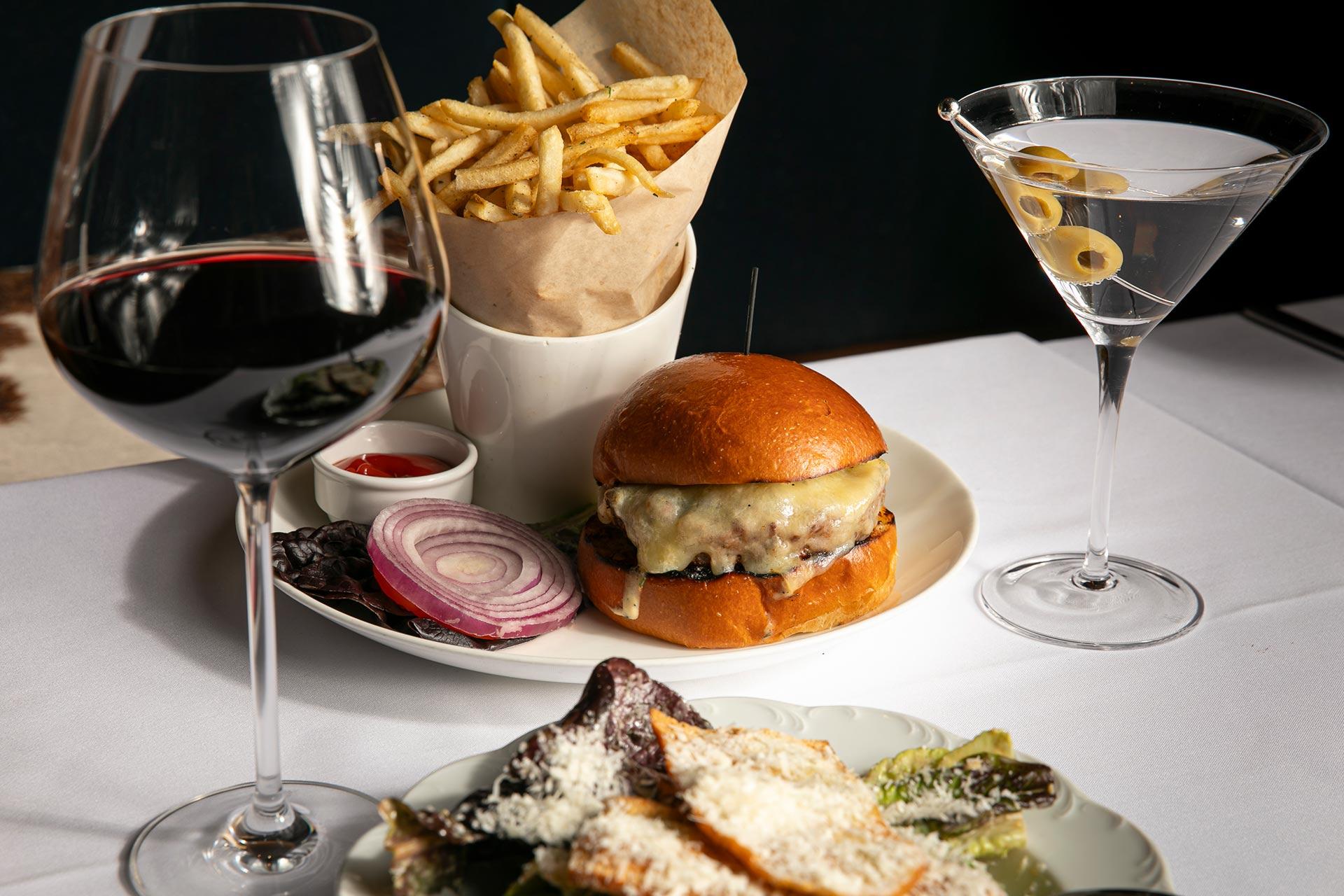 A cheeseburger with lettuce, tomato, and onion sits on a plate beside a serving of French fries and ketchup. A glass of red wine, a martini with olives, and a Caesar salad are also on the white tablecloth.