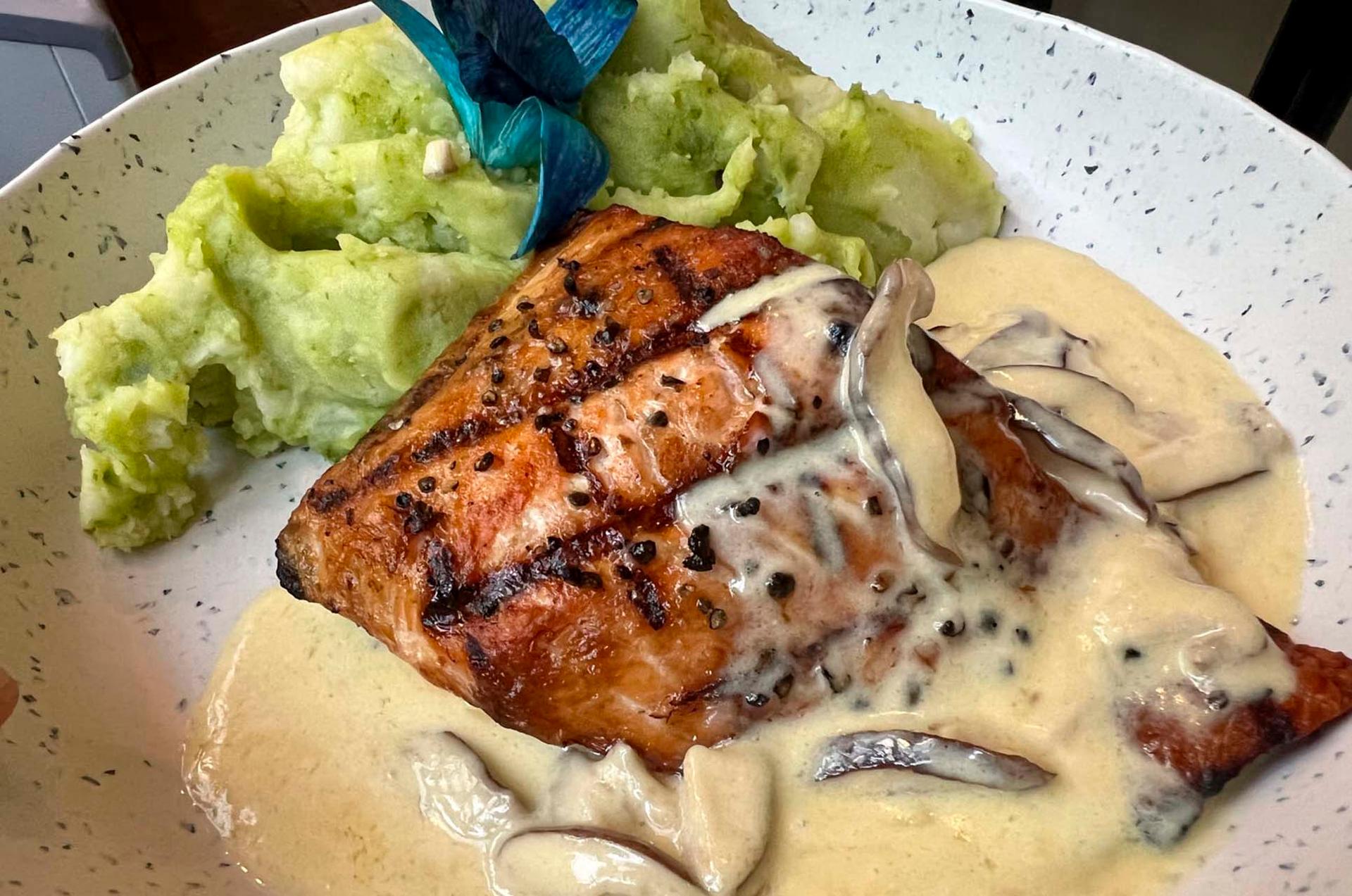 Grilled fillet with peppercorns, topped with creamy mushroom sauce, served with a side of green mashed potatoes and blue-tinted garnish on a speckled plate.