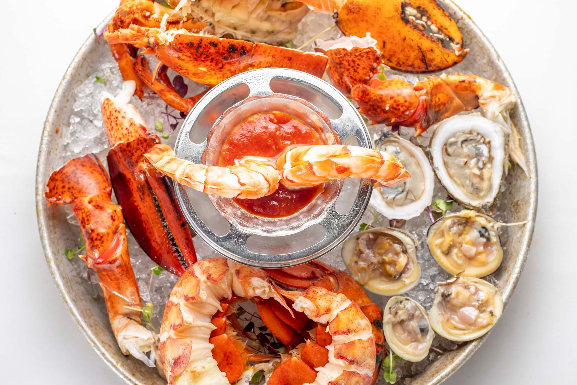 A seafood platter with lobster claws, shrimp around a cocktail sauce bowl, oysters, and clams on a bed of ice, served on a round metal tray.