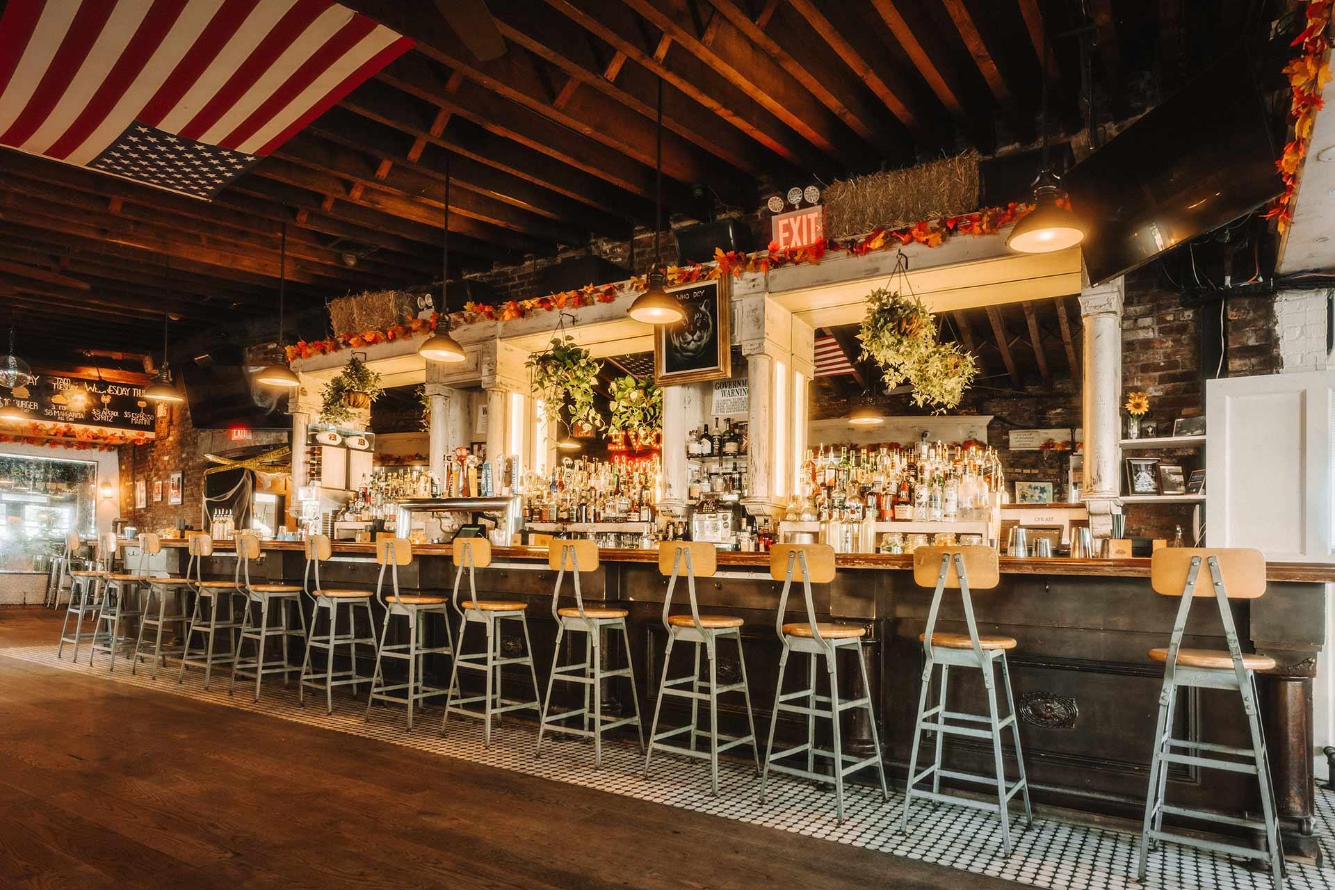 A rustic bar with a long wooden counter, metal barstools, hanging plants, shelves of bottles, exposed beams, and an American flag hanging from the ceiling. Warm lighting and autumn decorations create a cozy atmosphere.