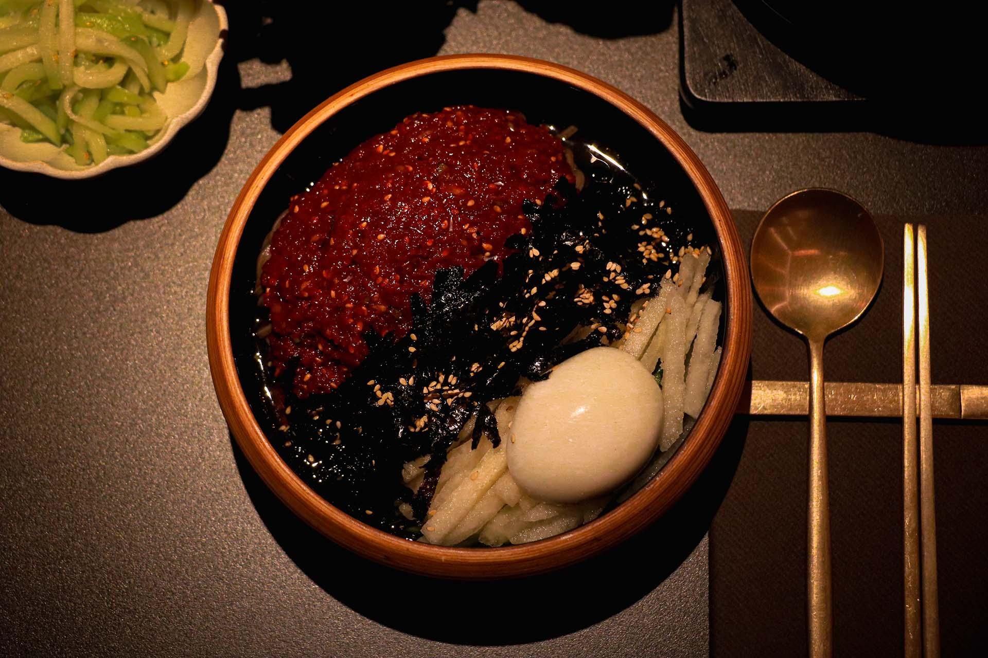 A wooden bowl with a spicy red sauce, seaweed, sesame seeds, shredded white veggies, and a boiled egg, next to a gold spoon and chopsticks. A small dish of green vegetables is partially visible on the side.
