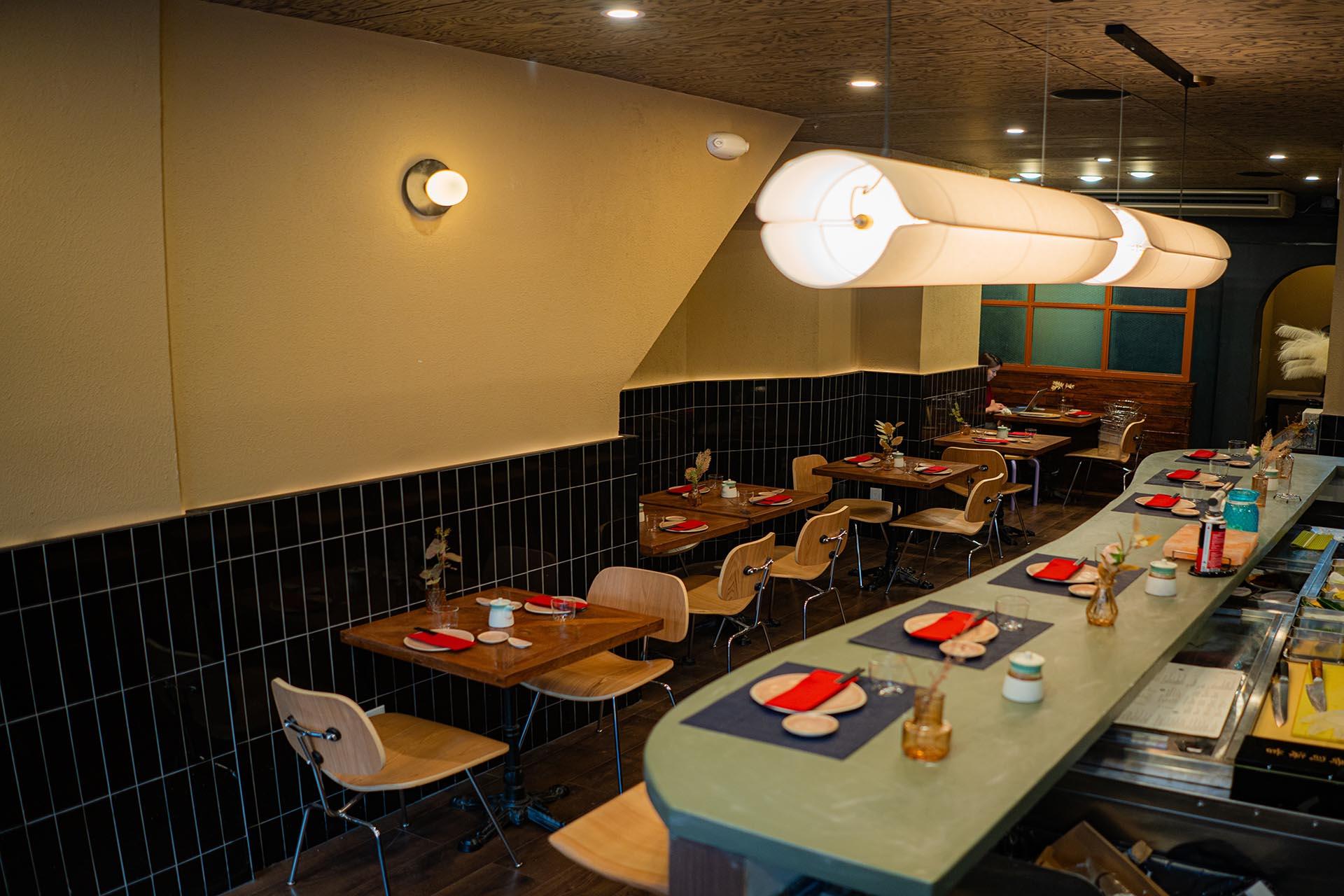 A modern restaurant interior with neatly set wooden tables, black tile walls, wooden chairs, and a sushi bar. Soft hanging lights brighten the cozy, inviting space.