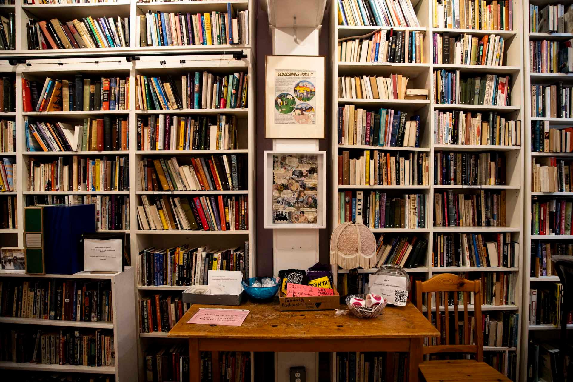 A wooden table with papers, bowls, and a fan sits in front of tall bookshelves filled with assorted books. Framed pictures and notes hang on the wall among the shelves. A wooden chair is beside the table.
