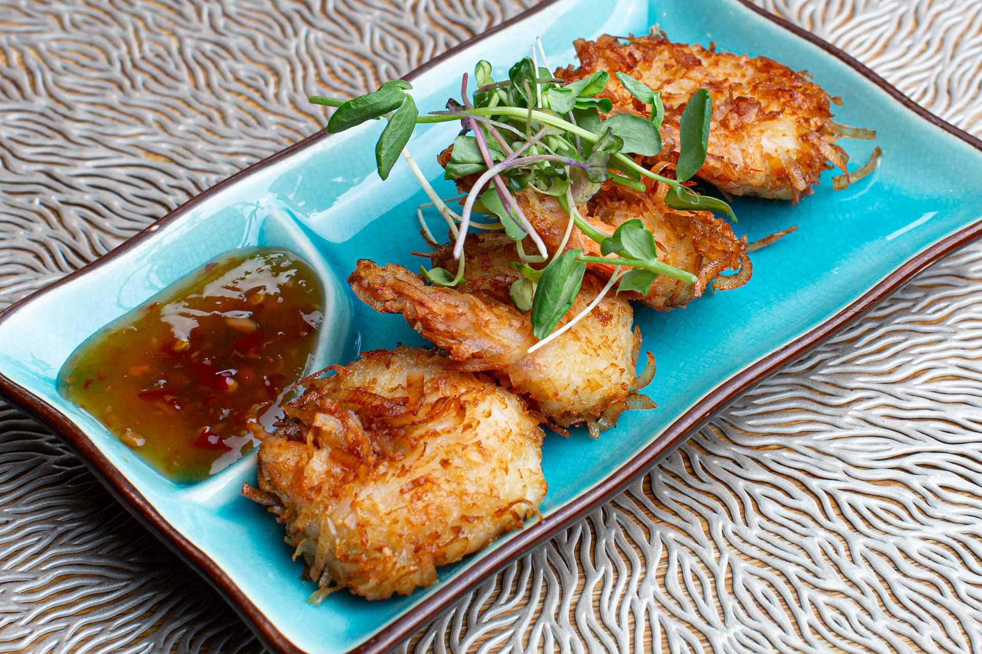Three crispy, golden-brown fritters garnished with fresh microgreens are served on a blue rectangular plate with a side of dipping sauce. The plate rests on a textured, wavy-patterned surface.