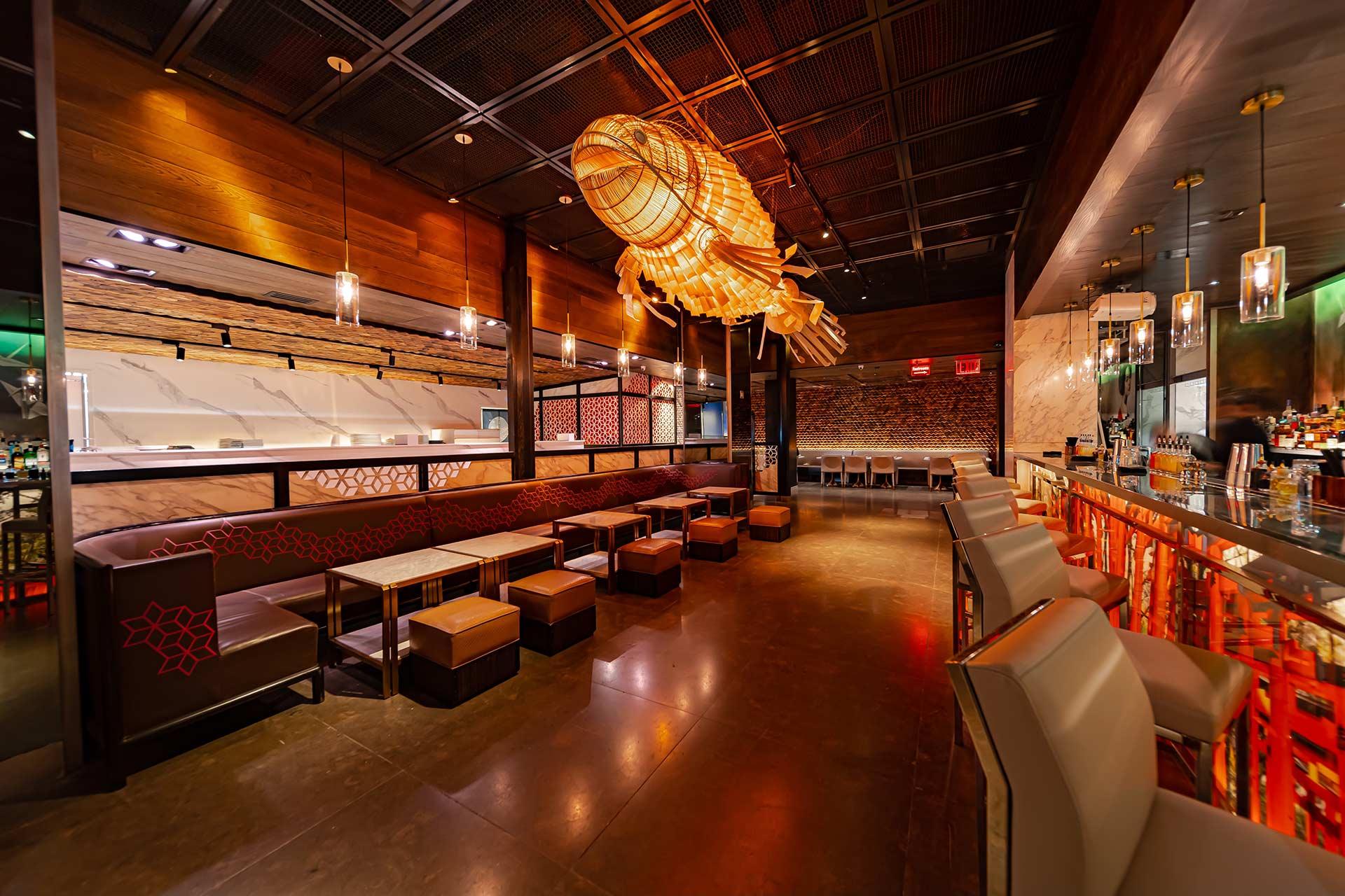 Modern restaurant interior with a large, illuminated fish-shaped light fixture, cushioned seating, a long bar with stools, and warm ambient lighting. The space features wood accents and a contemporary design.