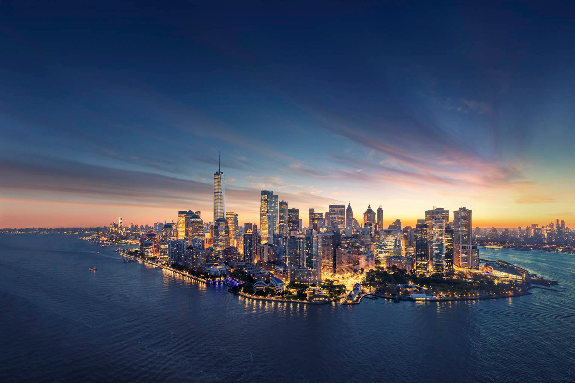 Aerial view of a city skyline at dusk, with skyscrapers illuminated and a mix of deep blue and orange hues in the sky. The water surrounding the city reflects the lights and colors, creating a serene and vibrant urban scene.