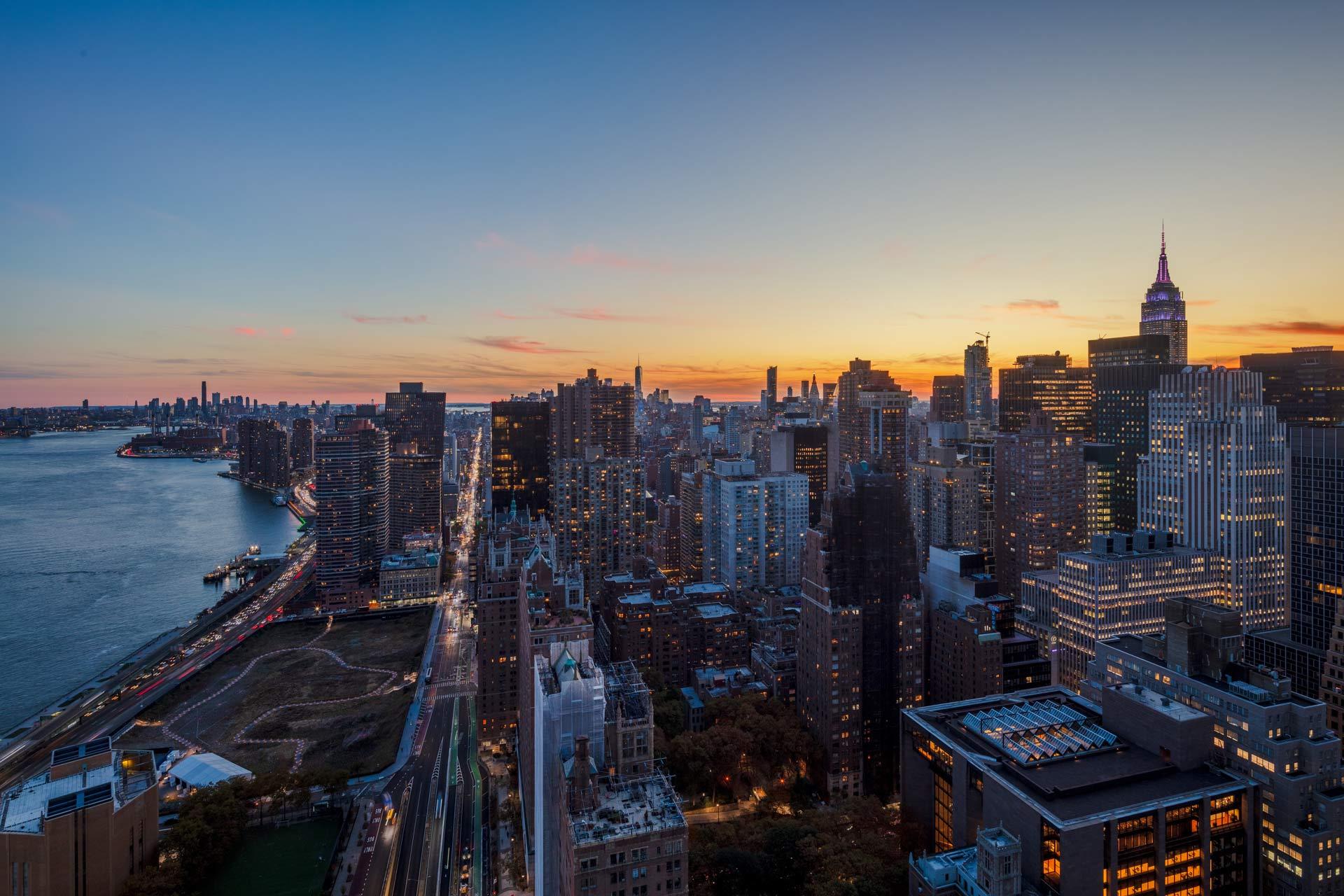 Aerial view of New York City at sunset, showing skyscrapers, the Empire State Building, and the East River with traffic along the adjacent roadway. The sky is tinged with orange and blue hues.