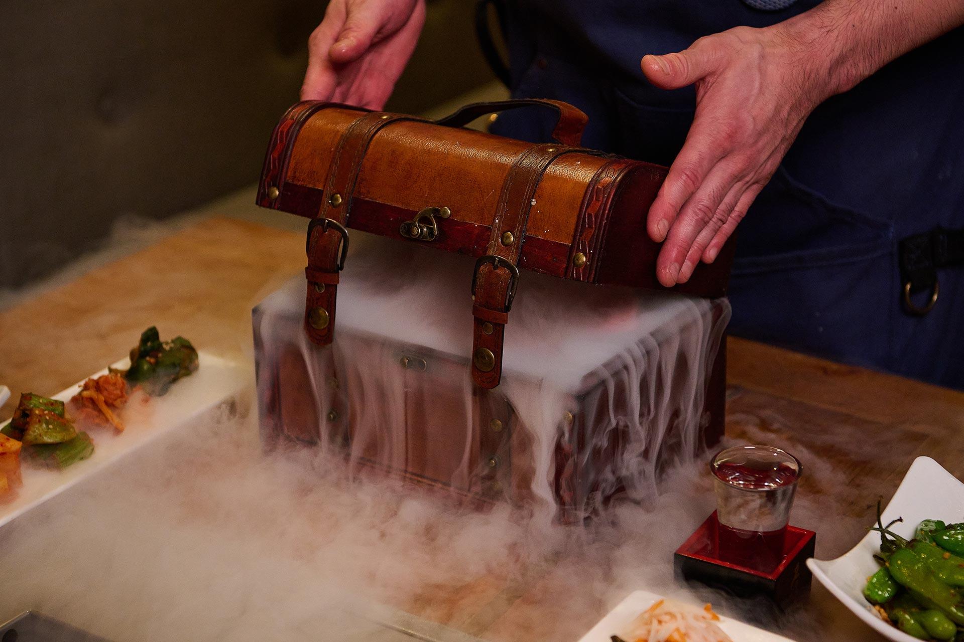 A person opens a vintage wooden chest with metal clasps, releasing dramatic white smoke that spills over the sides. Plates of food and a glass of liquid sit on the wooden table nearby.