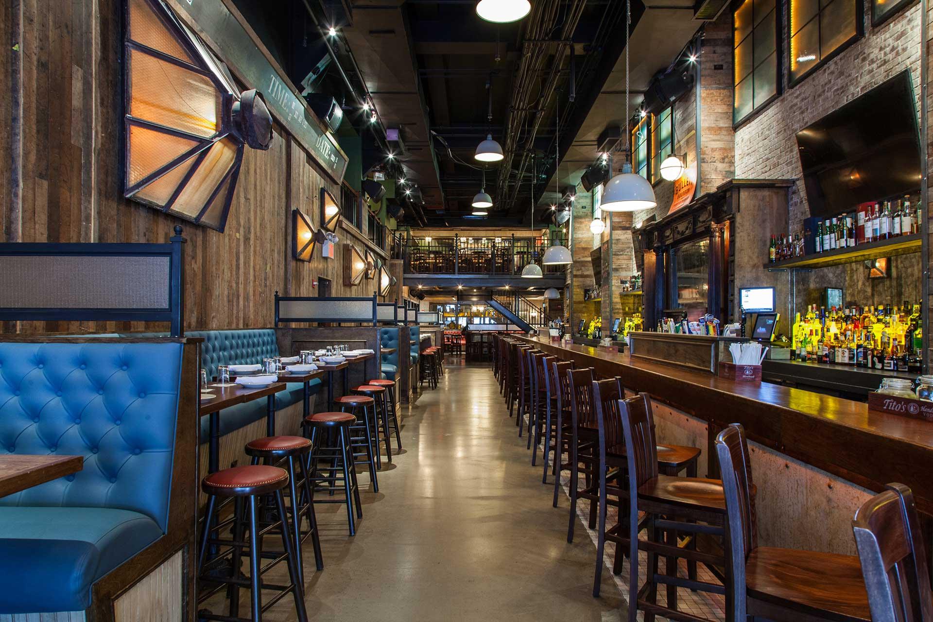 A rustic-style restaurant interior with wooden walls, high ceilings, a long bar with stools, blue upholstered booth seating, and tables set for dining; warm lighting creates an inviting atmosphere.