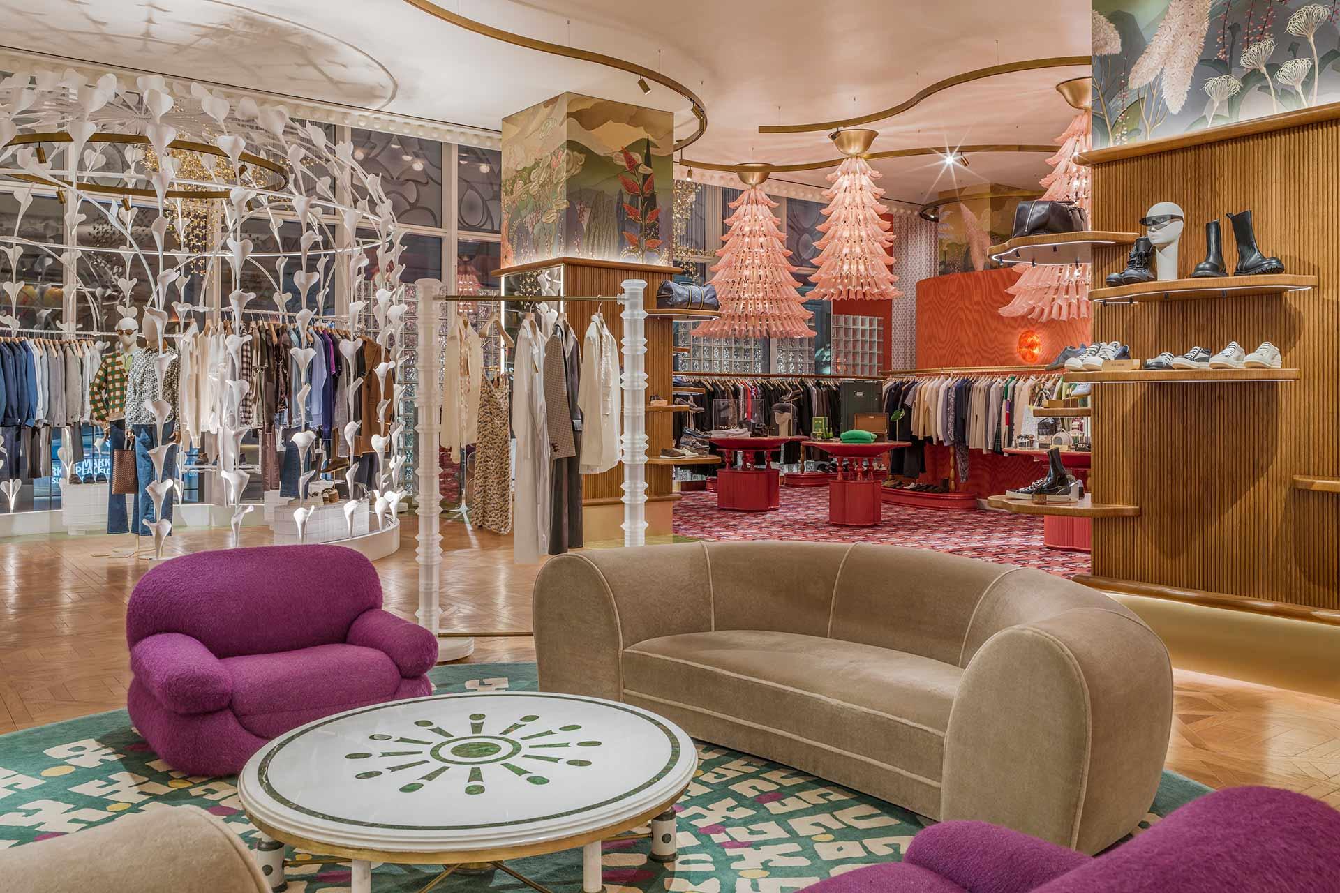 A stylish boutique with colorful decor, featuring purple armchairs, a beige curved sofa around a round table, racks of clothes, shelves with shoes, and unique hanging pink lamps.