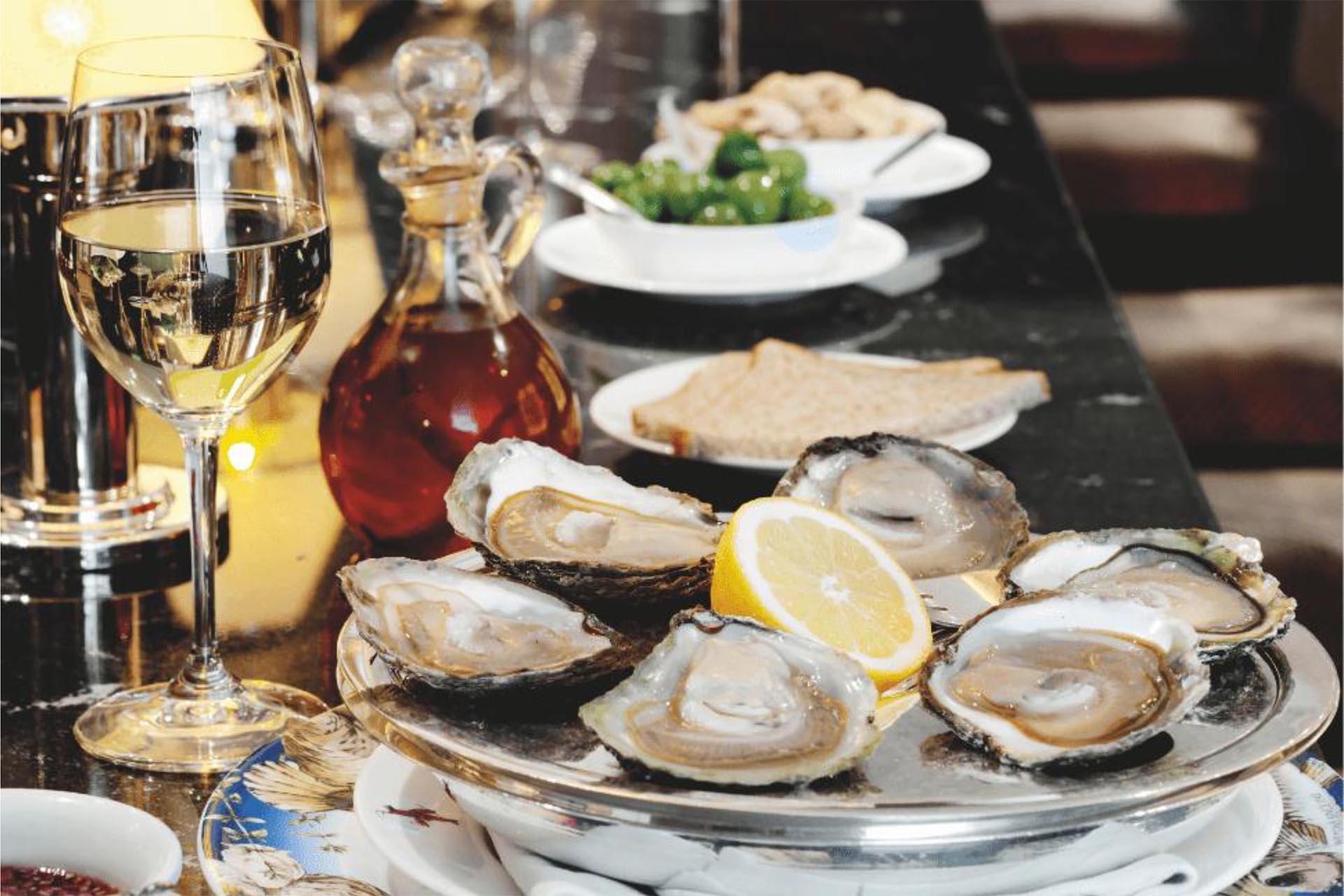 A plate of oysters with lemon wedges is on a table. Nearby, there's a glass of white wine, a small decanter, and plates with bread and green olives. The scene is set in a restaurant or dining setting.