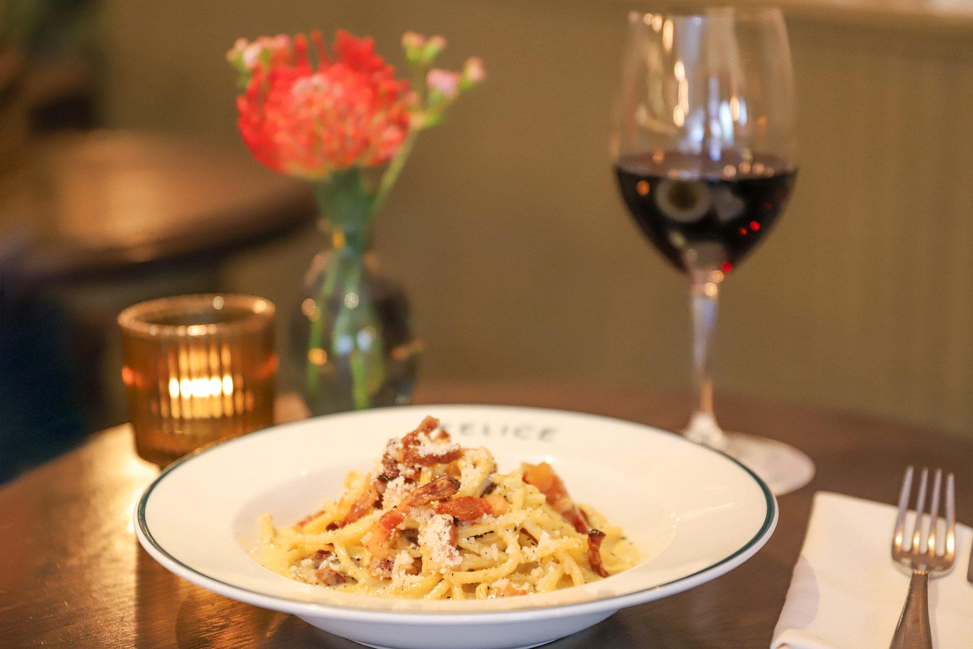 A plate of pasta topped with bacon and cheese sits on a table with a glass of red wine, a lit candle, a white napkin, and a small vase with red flowers in the background.