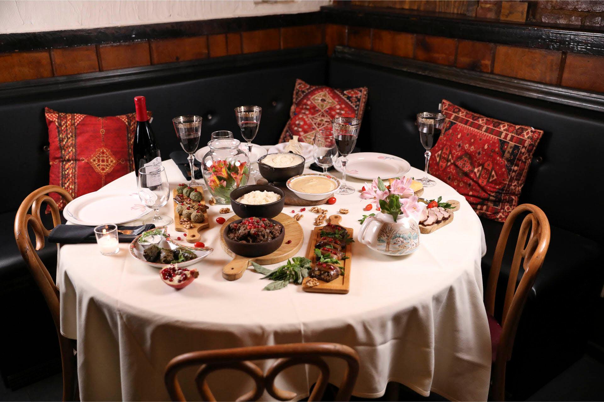 A round dining table set for four, featuring appetizers like sliced meat, bread, and dips. There are wine glasses, a bottle of red wine, and floral centerpieces. The seating includes cushioned benches with decorative red pillows.