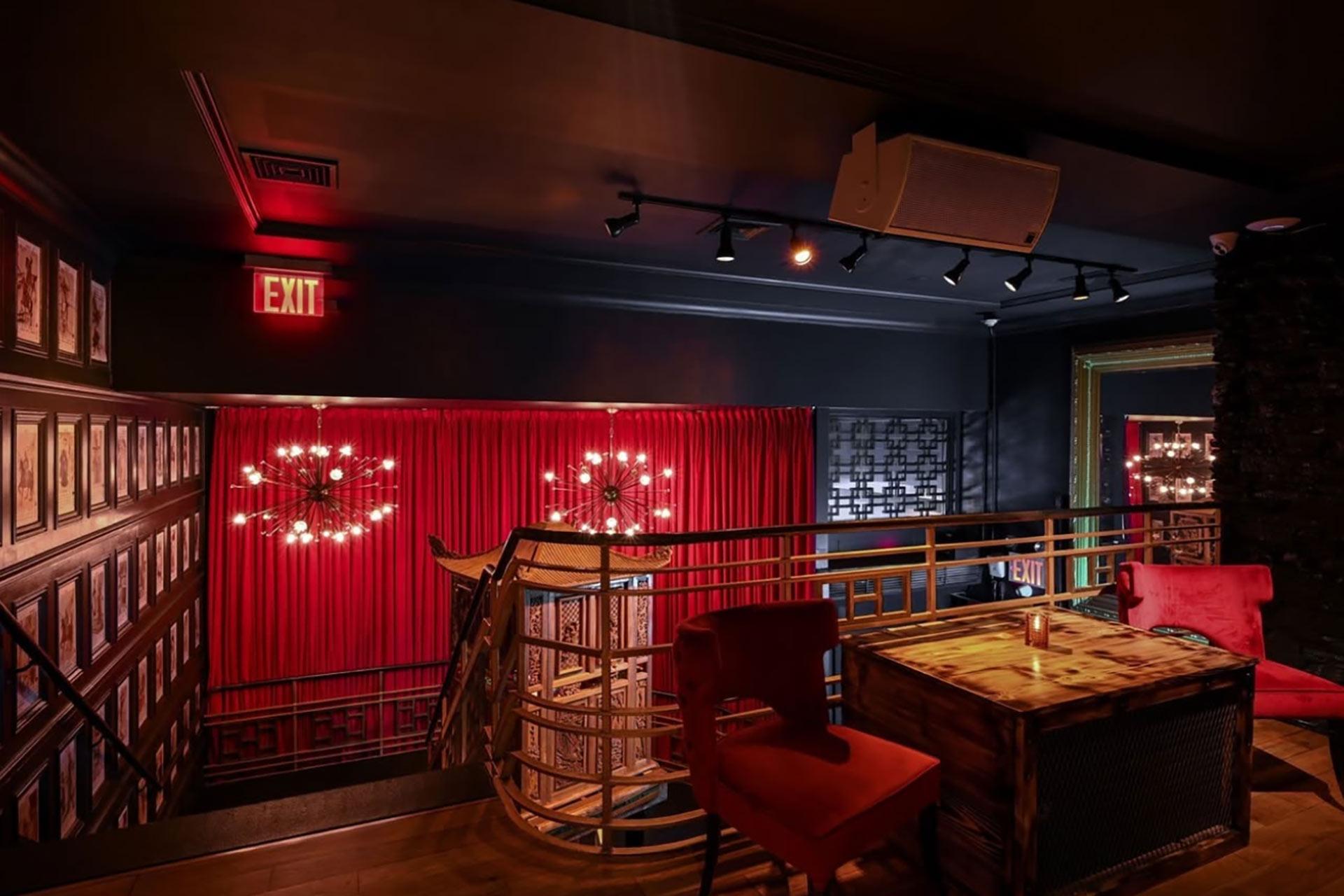 A dimly lit lounge with red curtains, red chairs, a wooden table, chandeliers, and framed photos on the wall. An EXIT sign glows above a stairway with a gold railing. The ambiance is cozy and intimate.