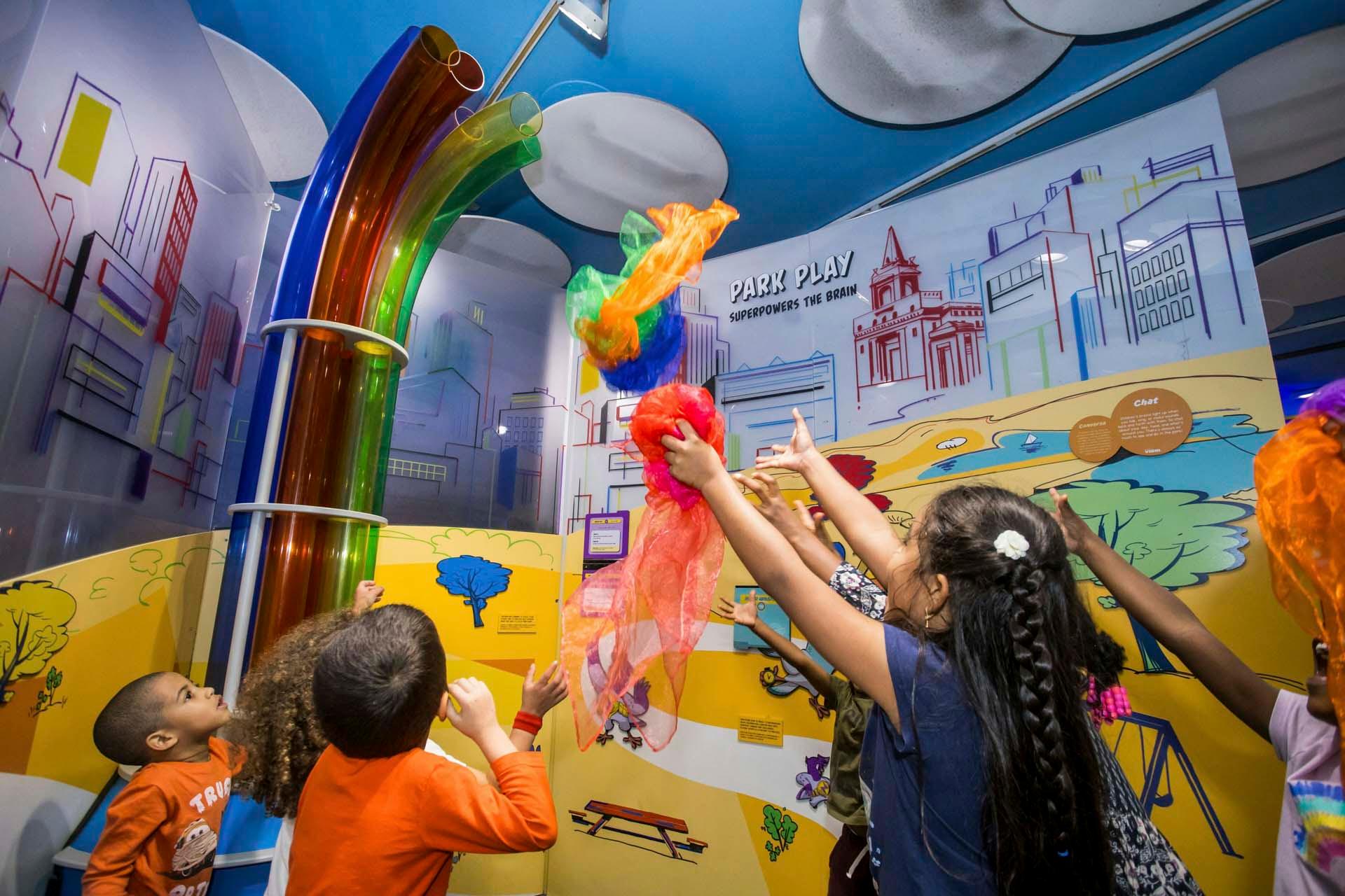 Children play indoors, tossing colorful scarves into the air near a display with curved transparent tubes and a mural of a city park scene on the wall behind them.