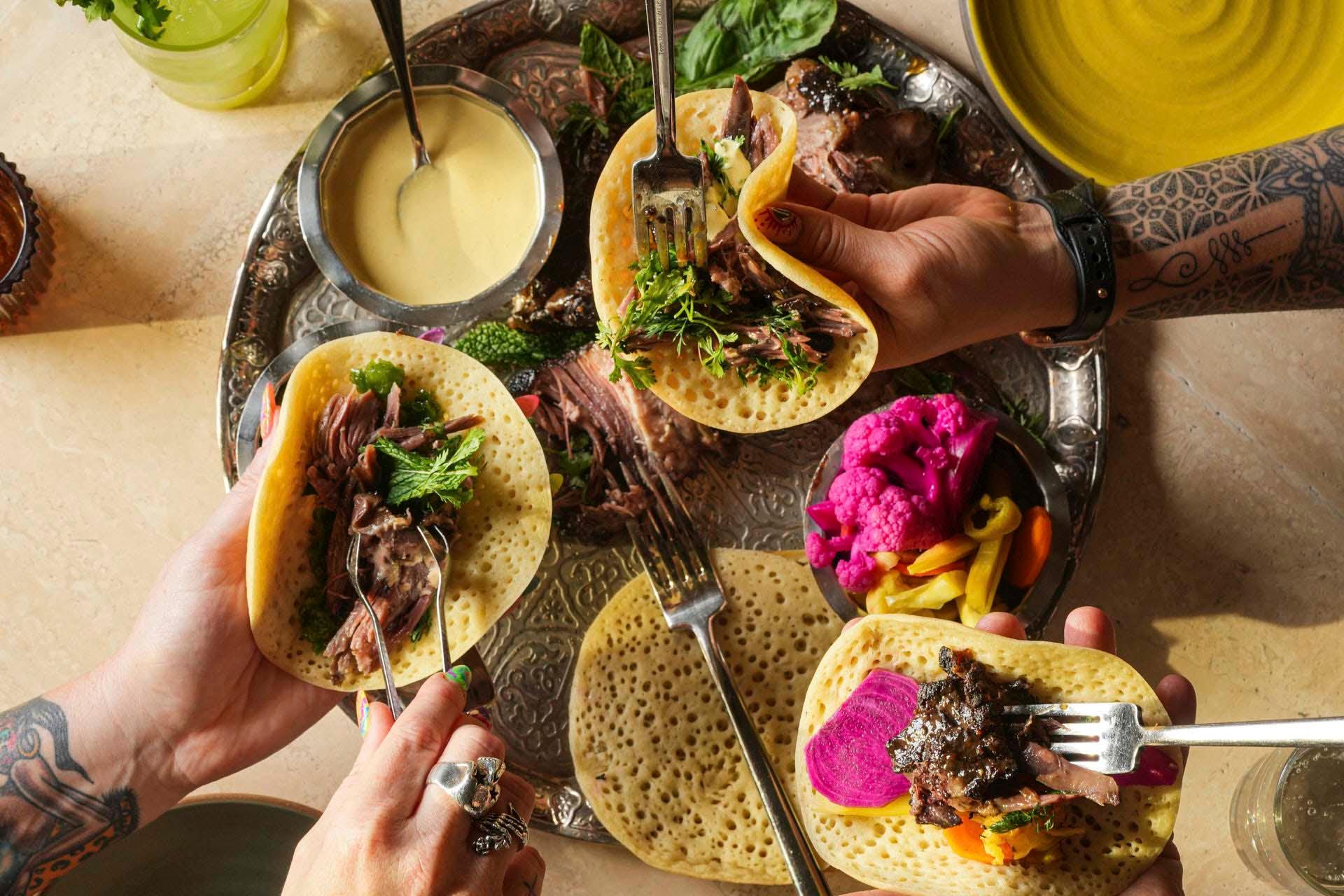 Three hands holding flatbreads filled with meat and herbs over a tray. The tray also holds pickled vegetables, and a bowl of dipping sauce is nearby. The table has plates and a drink, with tattoos visible on two wrists.