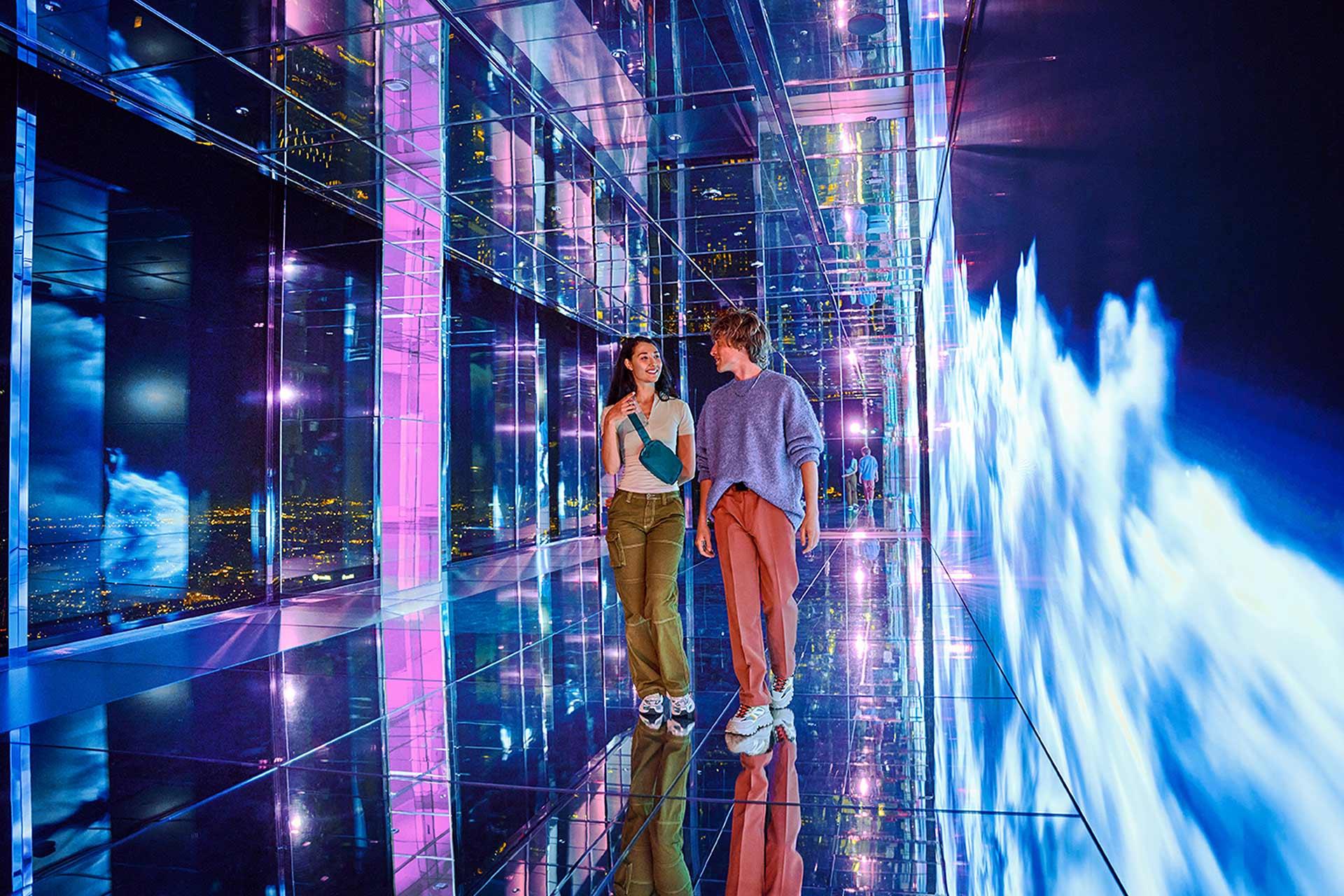 Two people walk and smile in a brightly lit, mirrored hallway with vibrant blue and purple lights and digital cloud-like displays on the walls and floor, creating a futuristic, immersive environment.