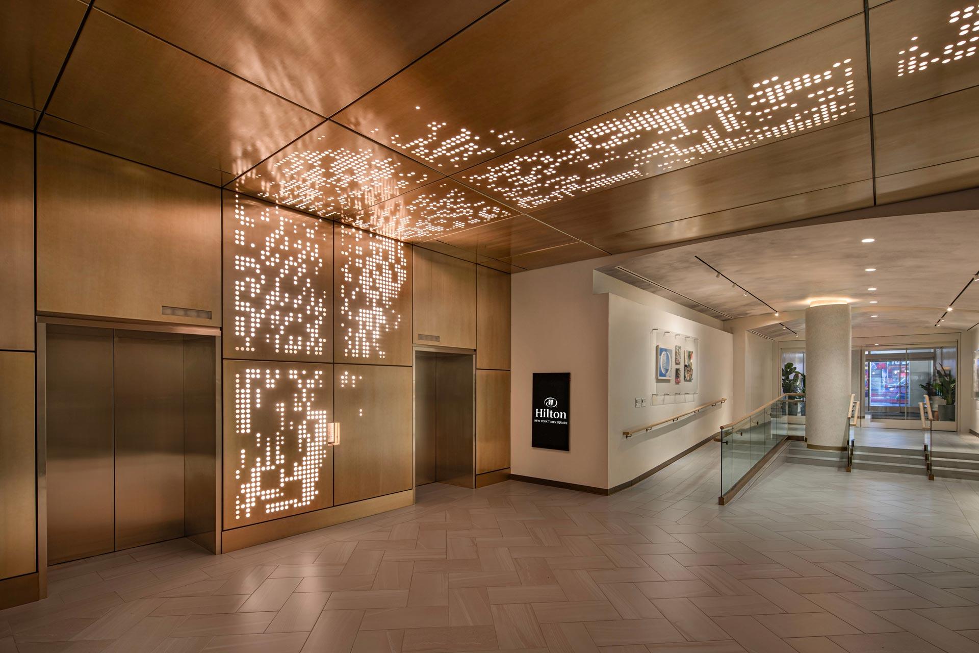 Hotel lobby with modern design featuring a wood-paneled ceiling and walls. An artistic light installation decorates the ceiling above two elevators. A sign with the Hilton logo is on the wall near a hallway leading further into the building.