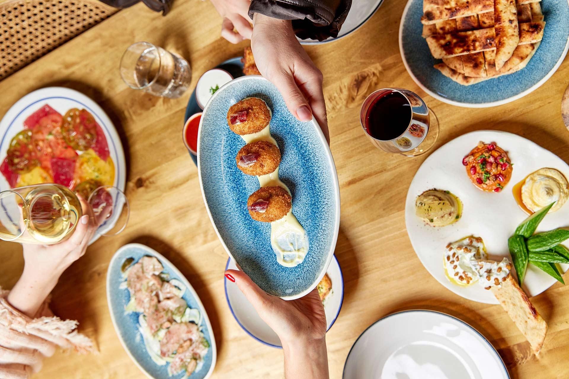 A hand passes a blue oval plate with three croquettes over a table set with various dishes, including bread, sliced tomatoes, appetizers, and drinks. Two people are reaching for food.