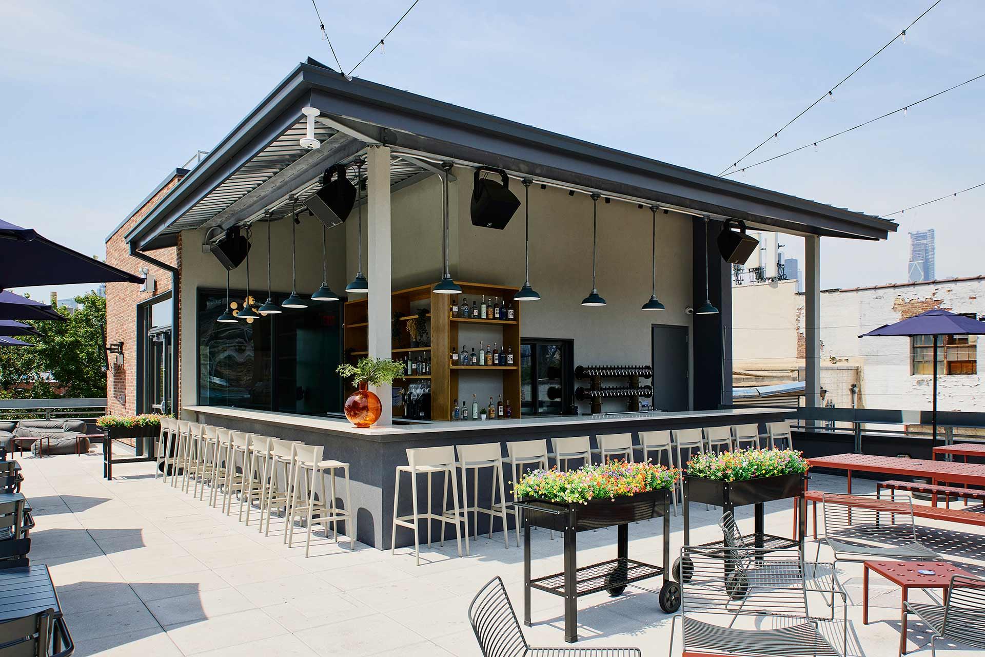 Modern rooftop bar with a central counter surrounded by barstools, hanging lights above, shelves stocked with bottles, planter boxes with flowers, string lights overhead, and outdoor tables and chairs under a clear sky.