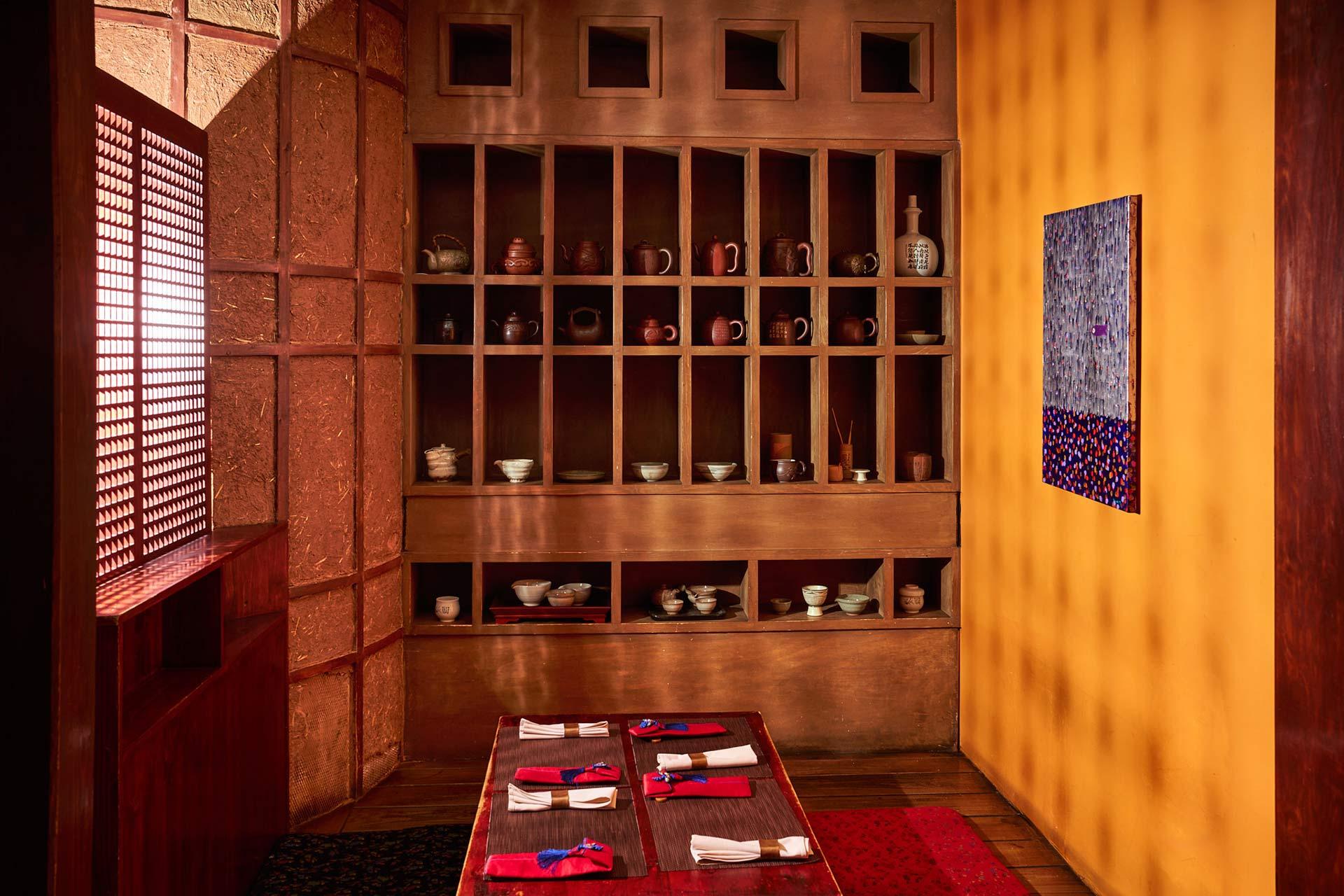 A cozy dining room with low wooden tables set with napkins and chopsticks, wooden shelves holding ceramic bowls, cups, and pots, and a yellow wall with abstract artwork. Warm lighting creates a welcoming atmosphere.