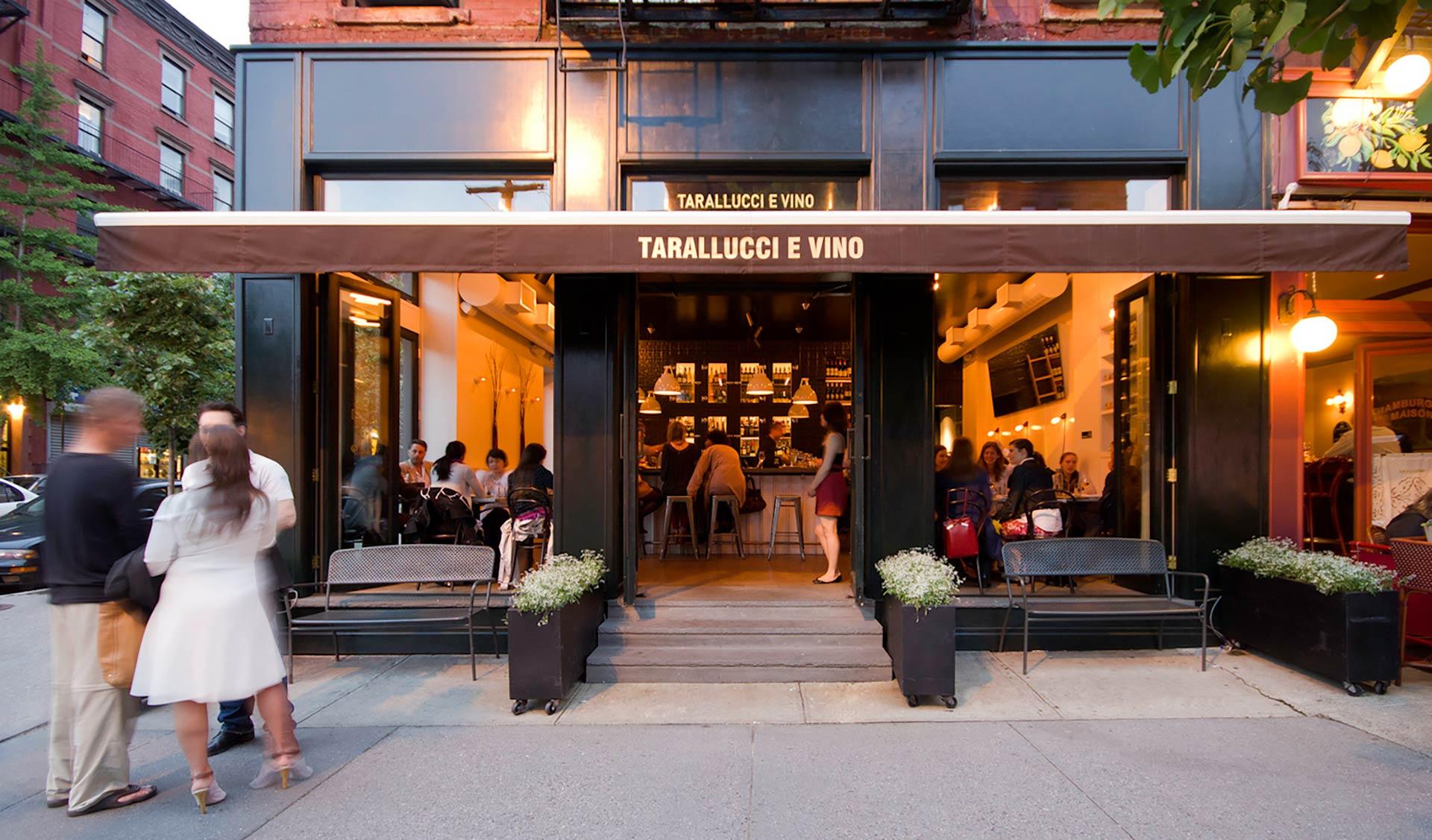People sit inside and outside a warmly lit restaurant called Tarallucci e Vino, with large windows, an awning, benches, and potted plants by the entrance on a lively city street.