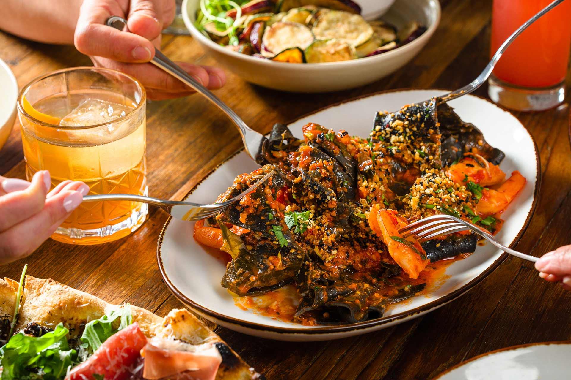 Three people share a plate of stuffed pasta topped with tomato sauce and herbs, surrounded by drinks, pizza, and a bowl of grilled vegetables on a wooden table.