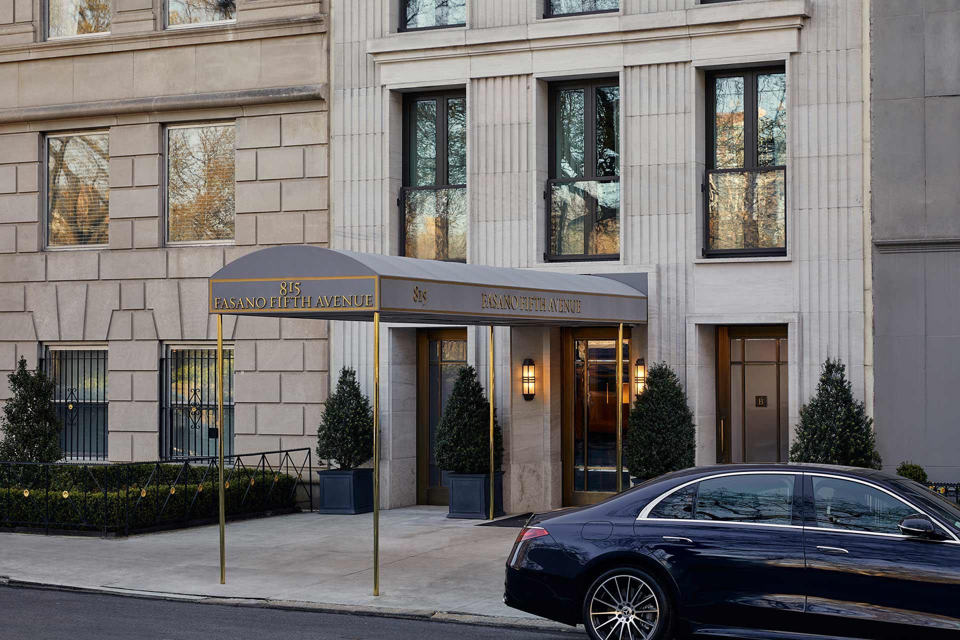 The entrance of a building with a gray canopy labeled "85 East 80th / 85 East 80th Avenue," flanked by potted plants. A black luxury car is parked on the street in front.