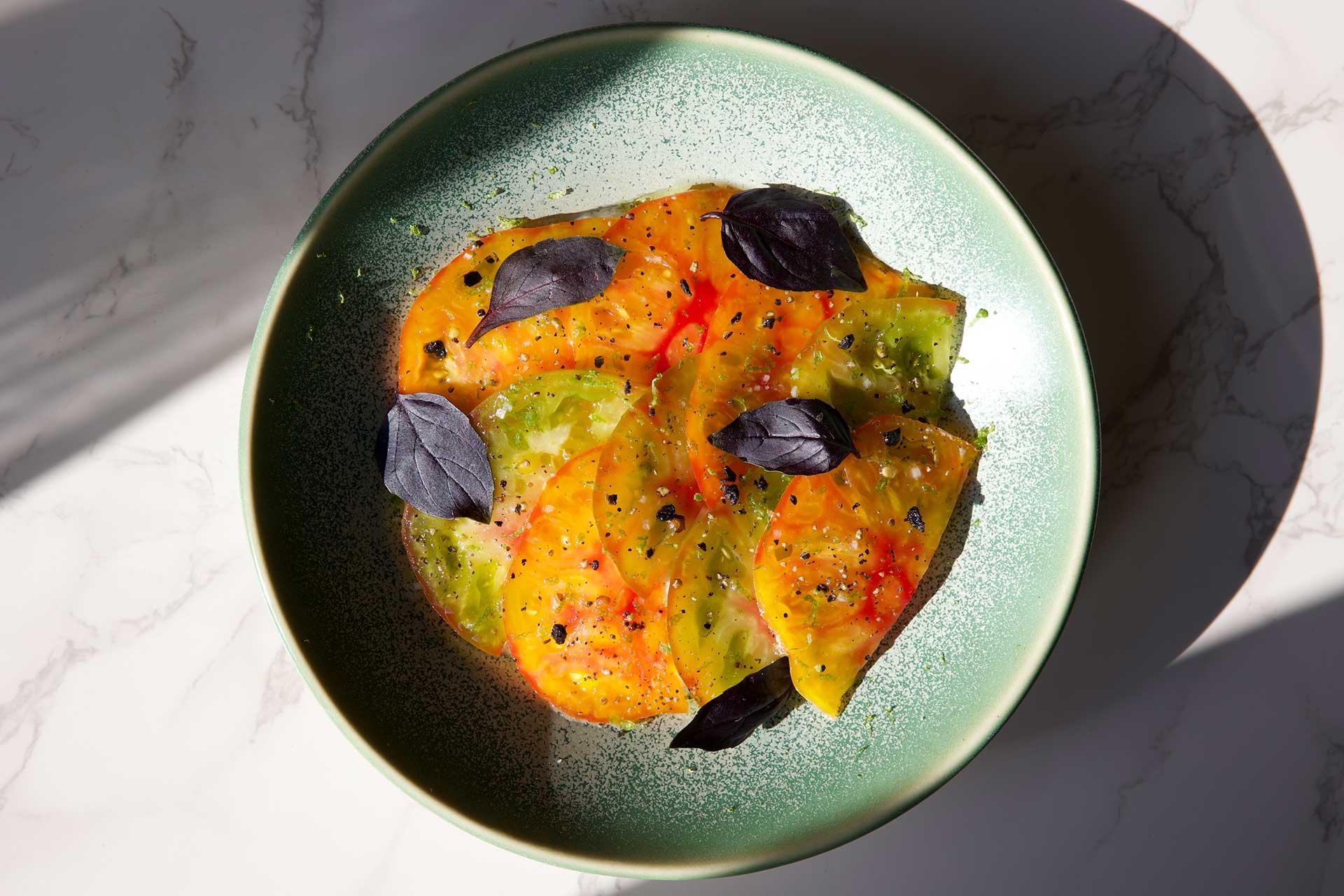 A green plate with thin slices of yellow and green heirloom tomatoes, garnished with black basil leaves, olive oil, and cracked black pepper, sitting on a marble surface in natural sunlight.