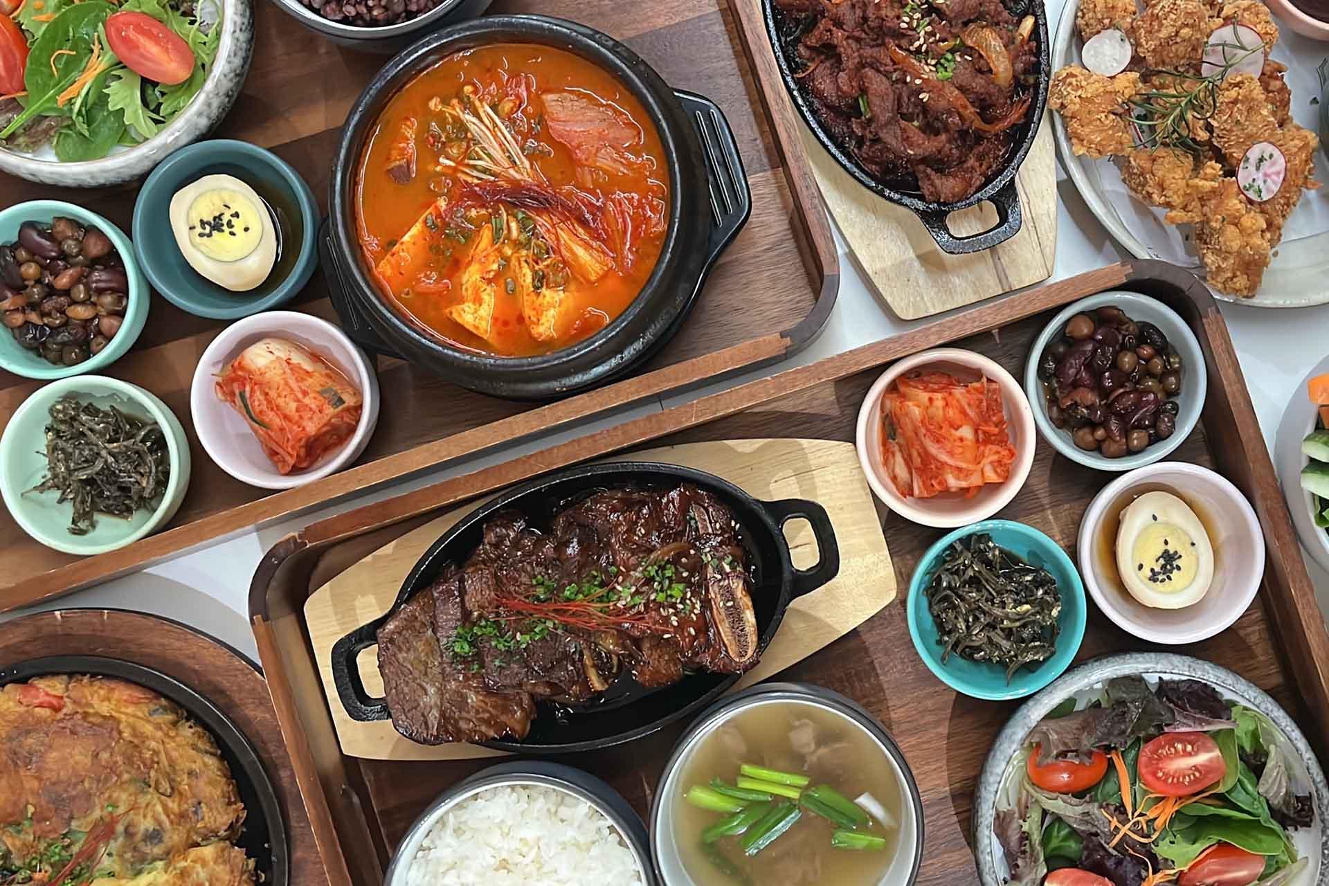 A table filled with various Korean dishes, including stews, grilled meat, fried chicken, kimchi, rice, salads, and assorted side dishes in bowls and plates.