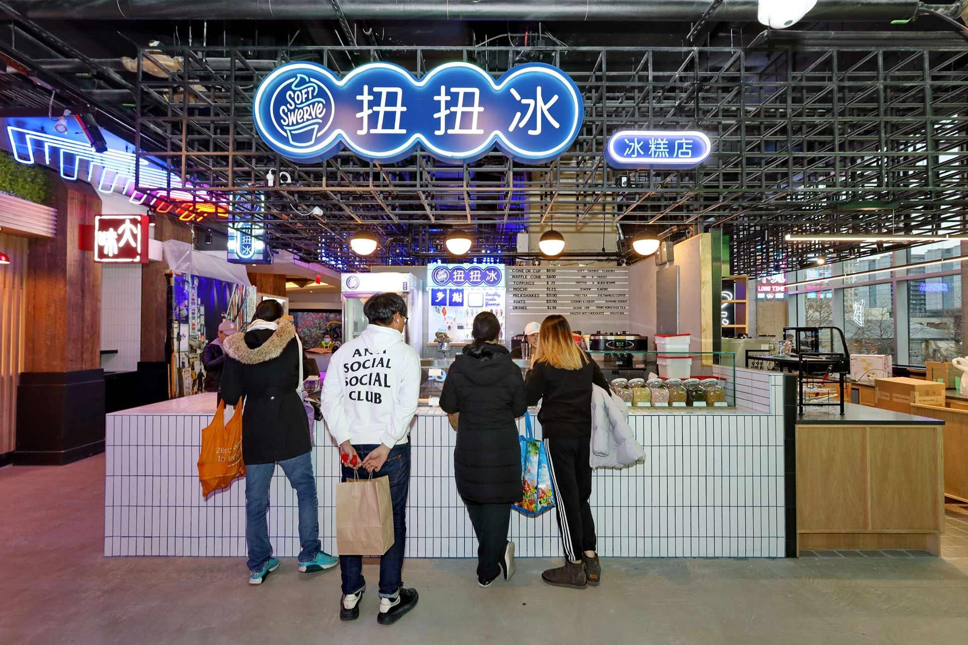 Four people stand at the counter of a brightly lit dessert shop with a blue neon sign displaying Chinese characters and "Soft Serve" in English. Various toppings are visible on the counter, and the shop has a modern design.