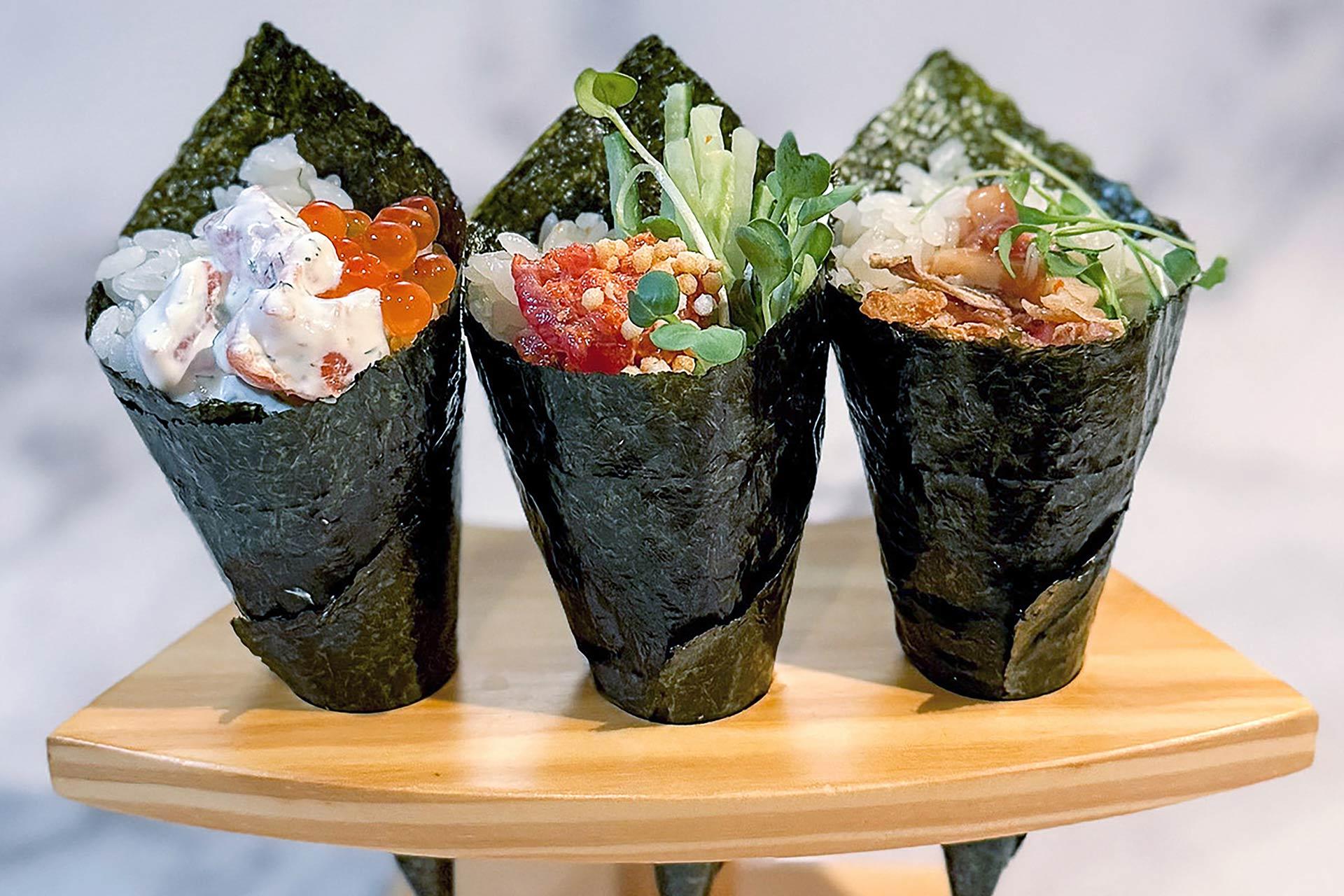 Three hand-rolled sushi cones (temaki) filled with various fresh ingredients including rice, fish, roe, and vegetables, are displayed upright on a wooden stand.