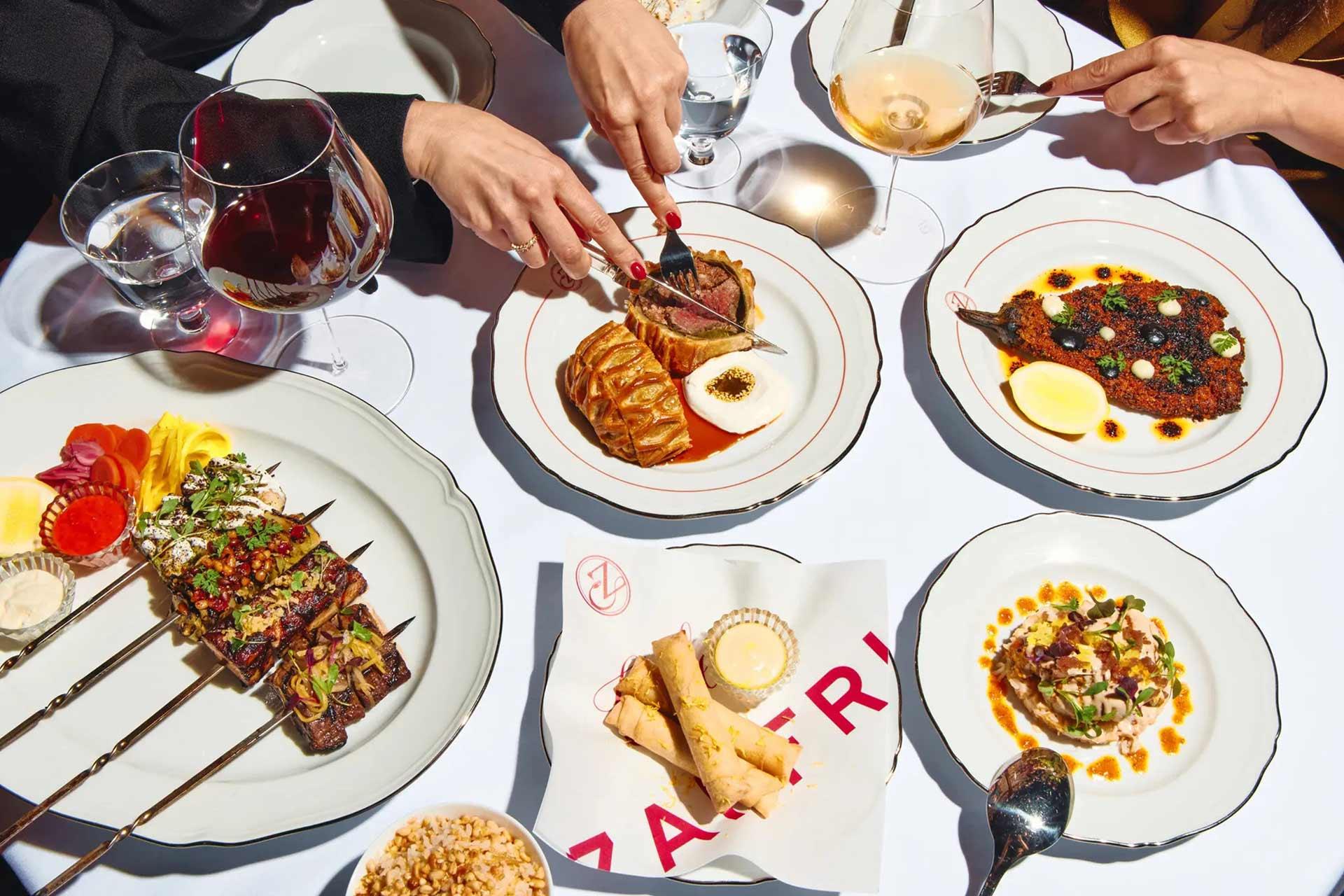 A table set with gourmet dishes and drinks, including skewers, meat with sauce, a plate of spring rolls, minced meat with eggs, and two glasses of wine, with two people’s hands serving and eating.