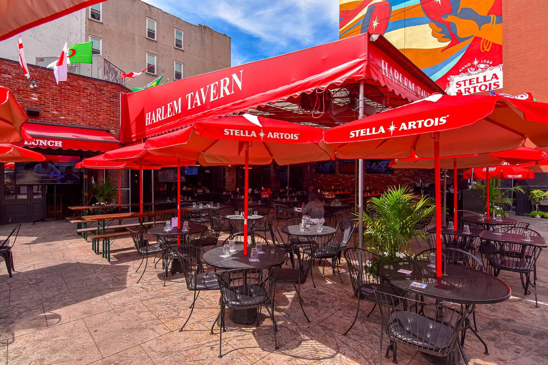 Outdoor seating area at Harlem Tavern with red Stella Artois umbrellas, metal tables and chairs, potted plants, and a colorful mural on a nearby building wall under a bright blue sky.