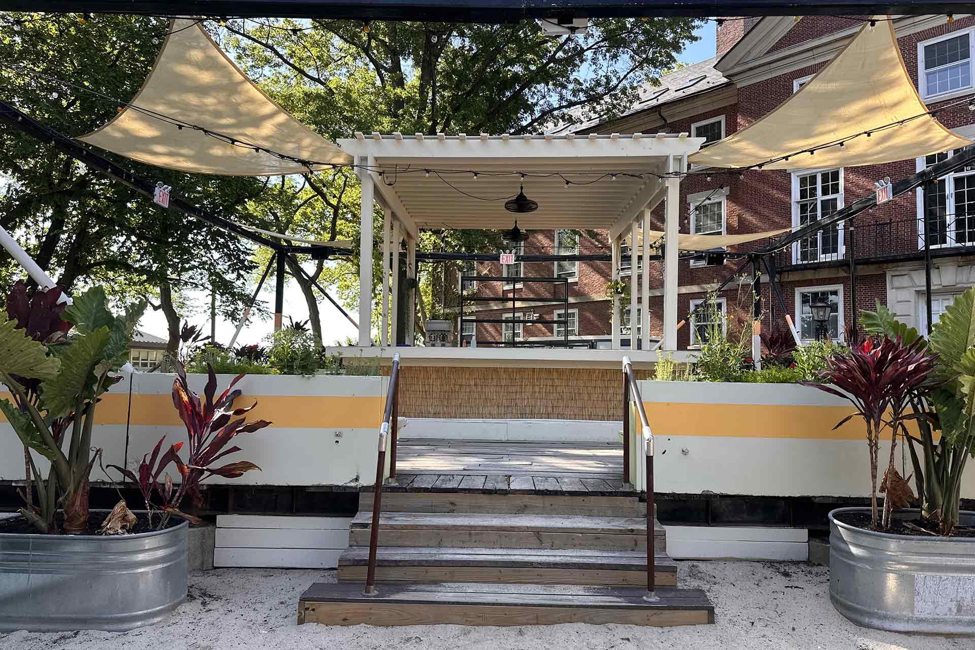 Outdoor patio with canopy sails, wooden steps, and metal railings, surrounded by tropical plants in metal containers. In the background, a brick building with windows is partially visible through trees.