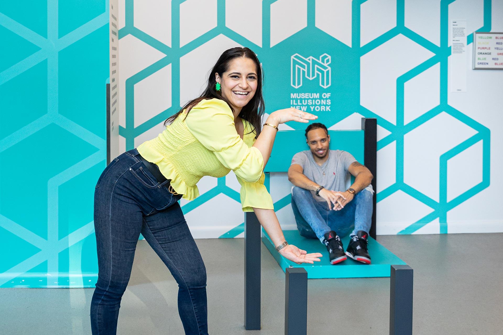 A woman poses, appearing to hold a tiny man on her hand due to an optical illusion with a giant chair at the Museum of Illusions New York. The background is teal with geometric patterns.
