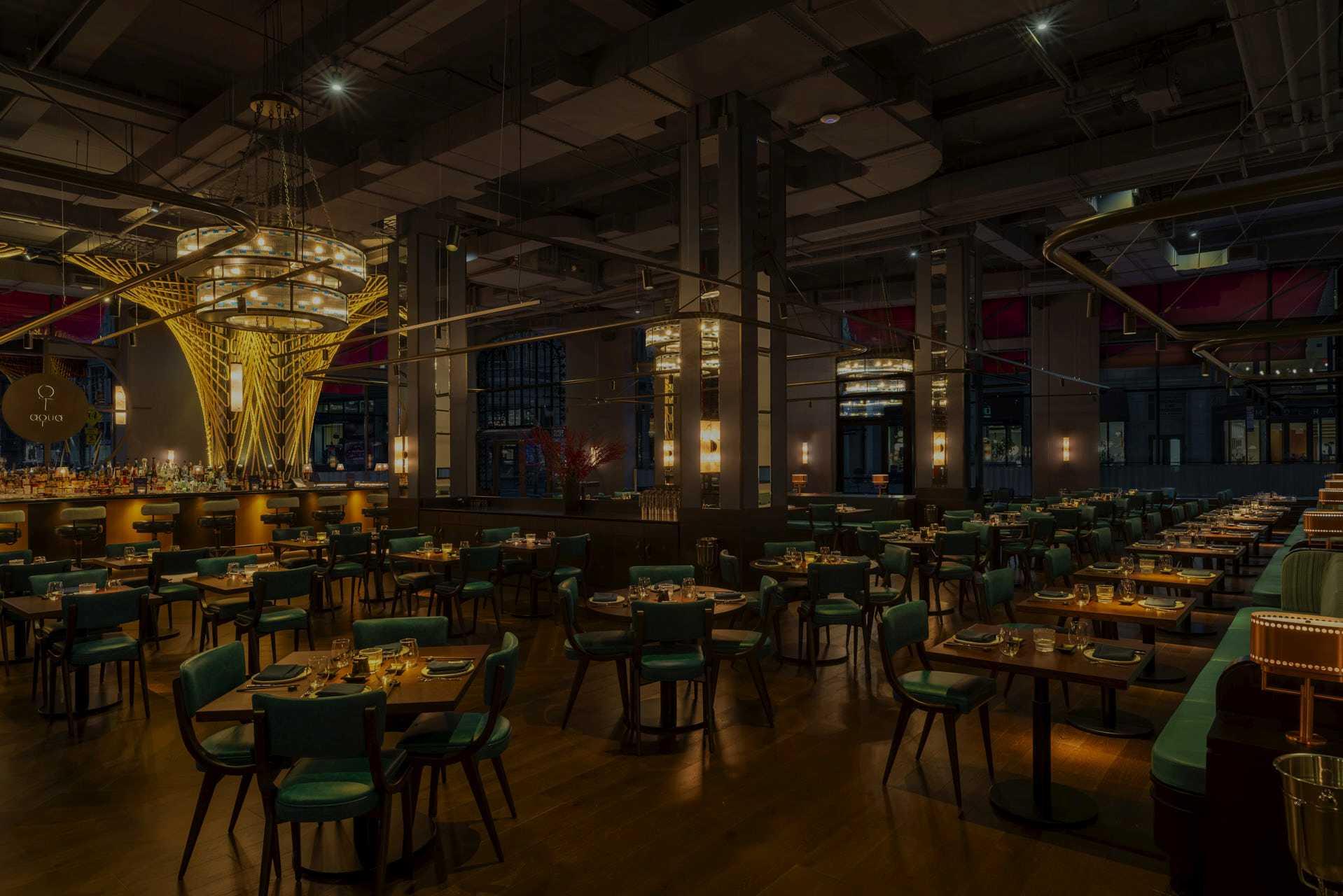 Spacious, modern restaurant interior with dim lighting. Wooden tables and chairs fill the room, with settings for dining. A well-lit bar with tall, artistic light fixtures is in the background. The atmosphere is warm and inviting.