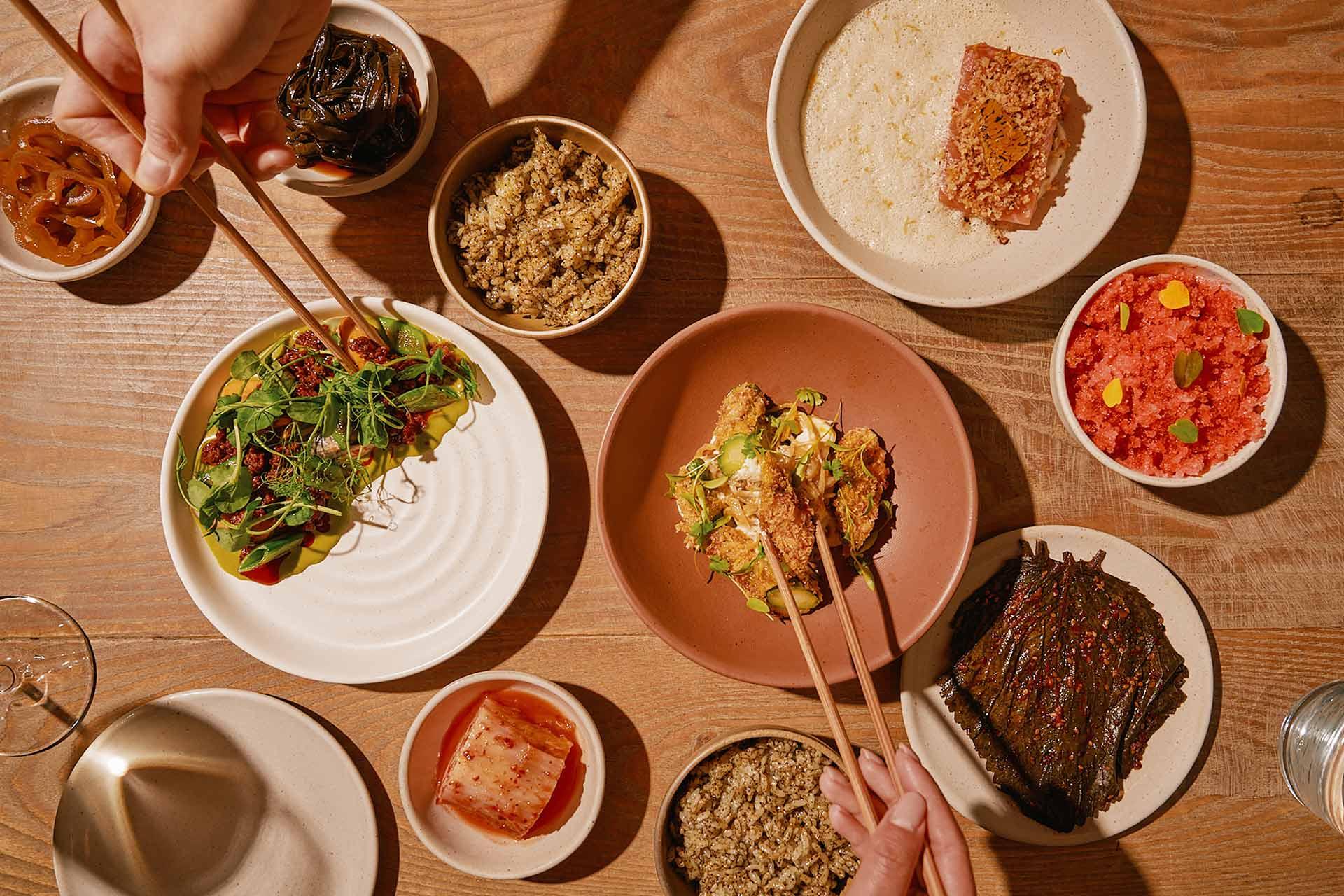 Overhead view of a wooden table set with various small dishes of food, including rice, vegetables, pickles, and fish. Two pairs of hands use chopsticks to pick up food from different plates.