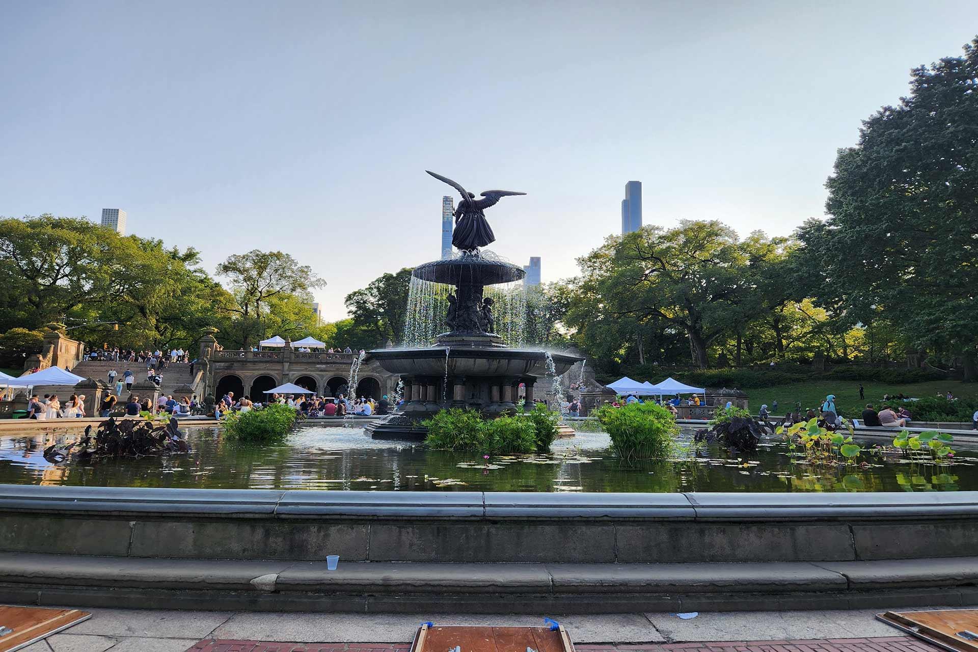 A large fountain with an angel statue stands in the center of a pond in a park, surrounded by trees and people. City buildings are visible in the background under a clear sky.