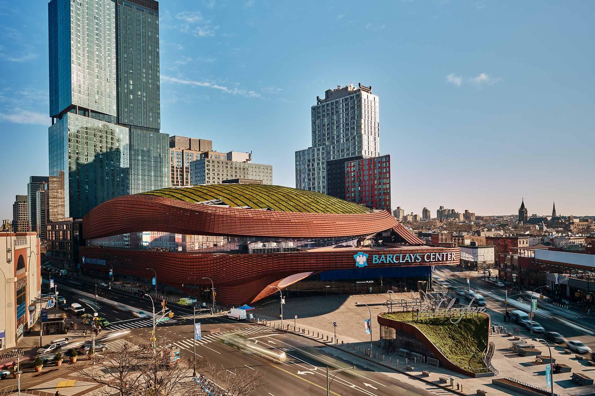 A wide view of the Barclays Center in Brooklyn, New York, showing its distinctive brown, curved design with a green roof, surrounded by city streets and tall buildings under a clear blue sky.