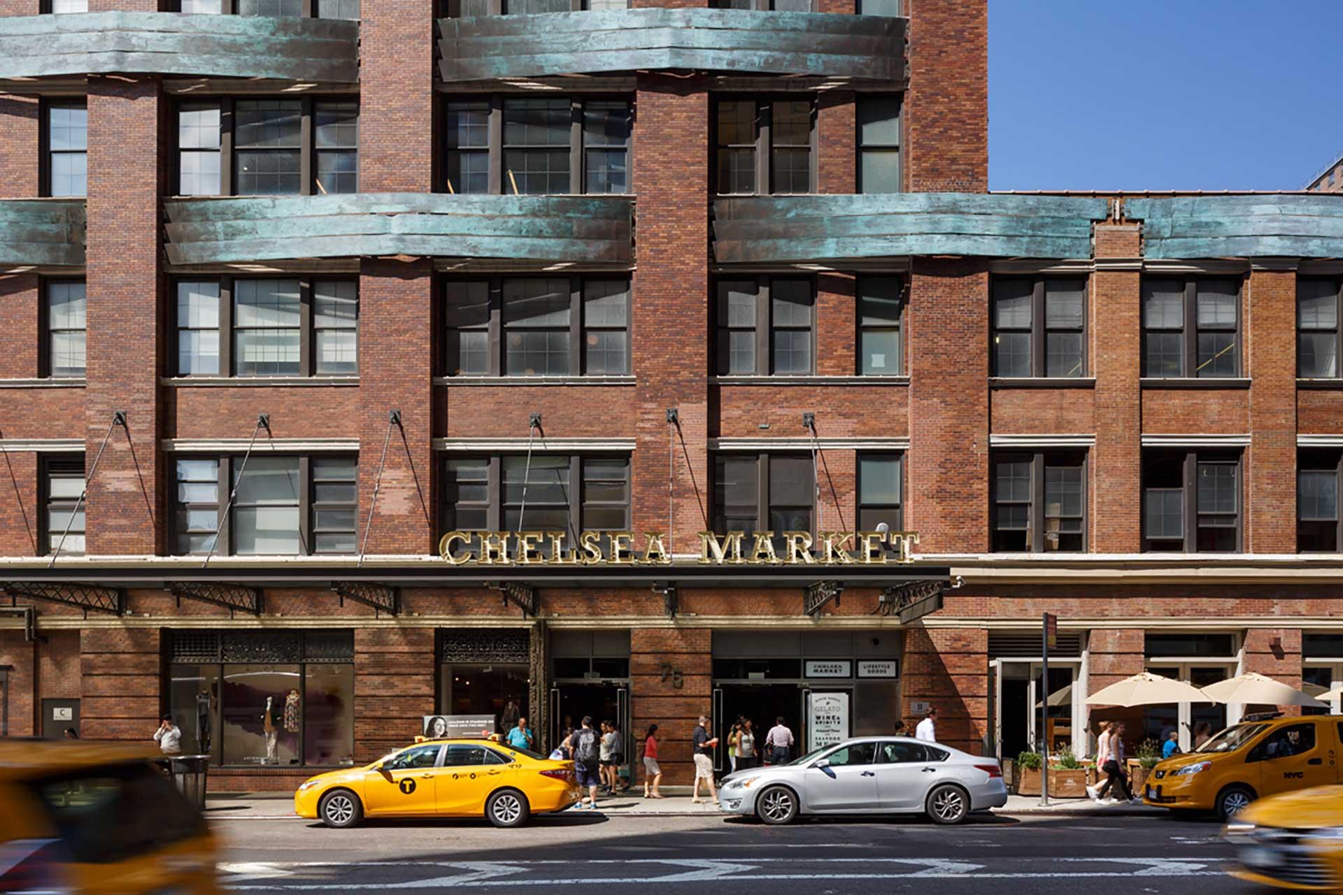 Street view of the Chelsea Market building with large windows and a sign above the entrance. Yellow taxis and other cars are passing by, and people are near the entrance and outdoor seating area.