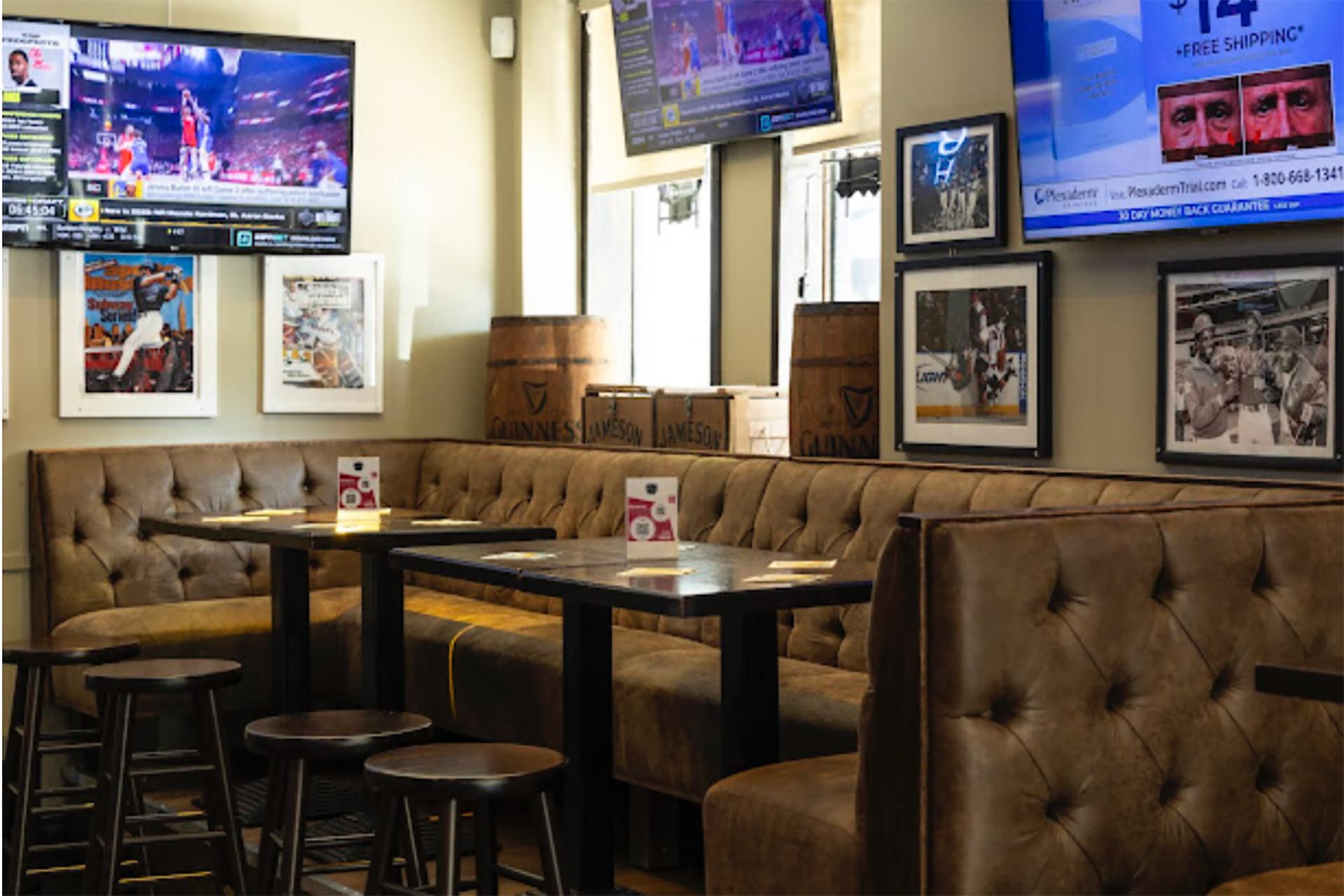 A cozy sports bar with brown tufted booths, round wooden stools, and tables. Multiple TVs display sports and ads. Framed sports photos and wooden crates decorate the walls. Natural light streams in through the windows.